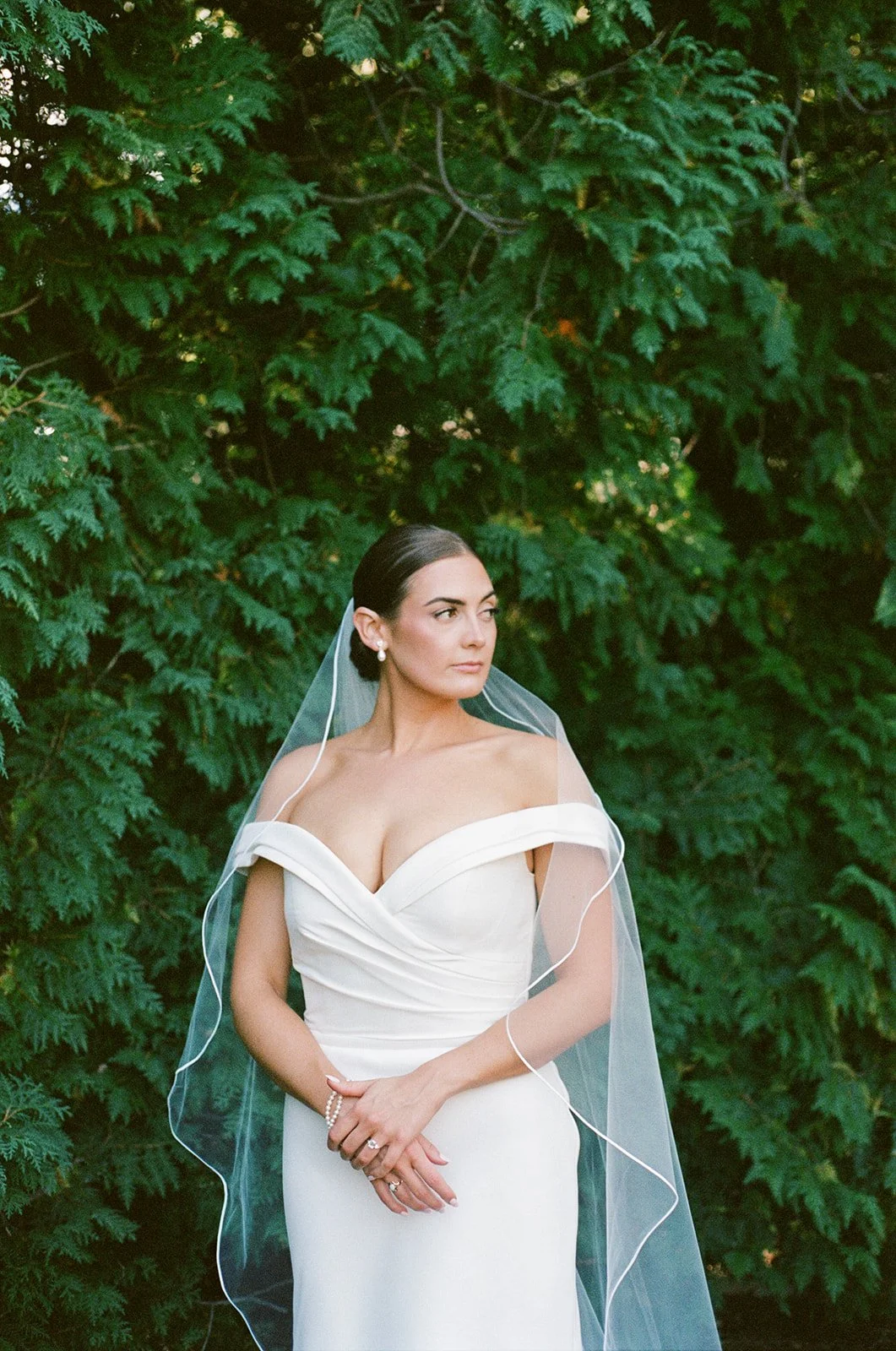 Brunette bride in a white off-the-shoulder wedding dress with a veil, standing outdoors in front of green foliage, looking to her left.