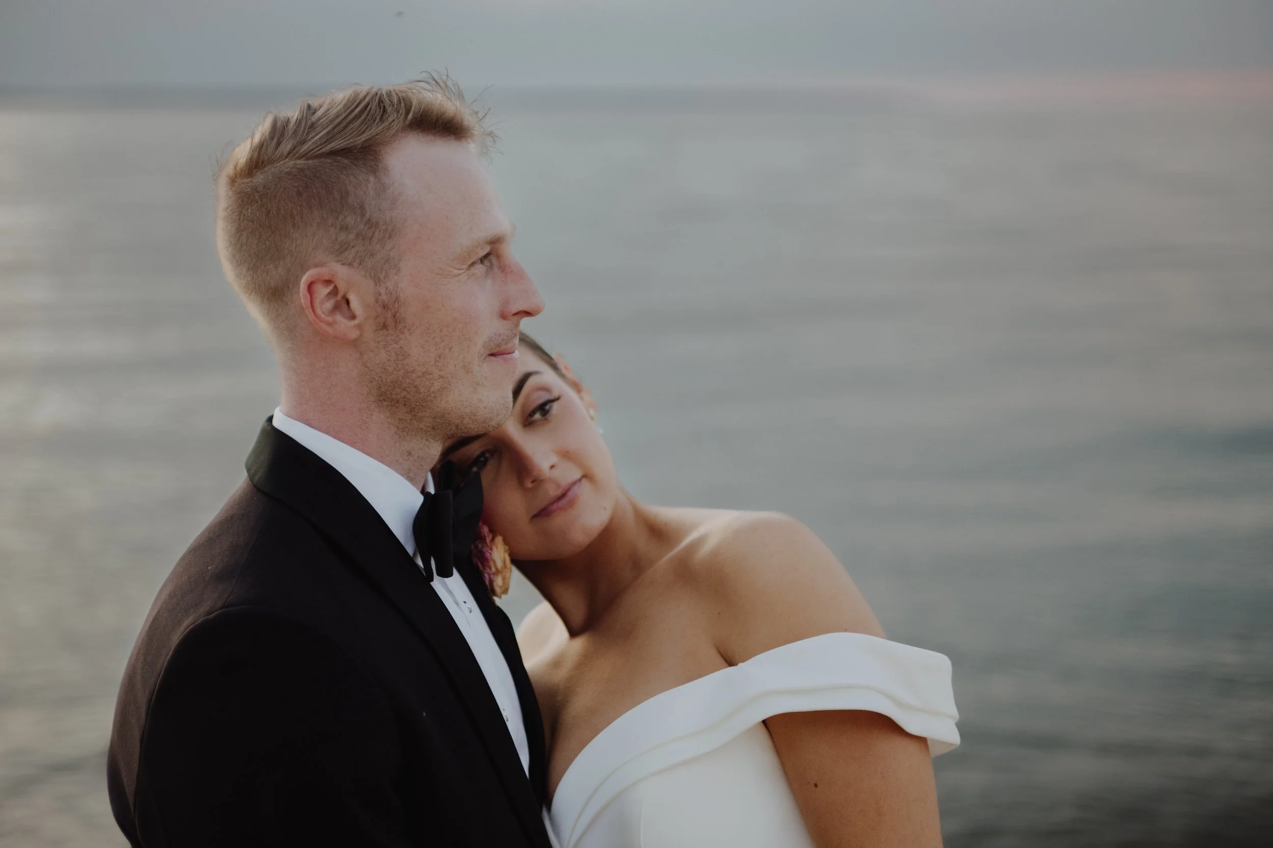 A bride and groom embrace by the water during sunset, with the bride resting her head on the groom's shoulder.