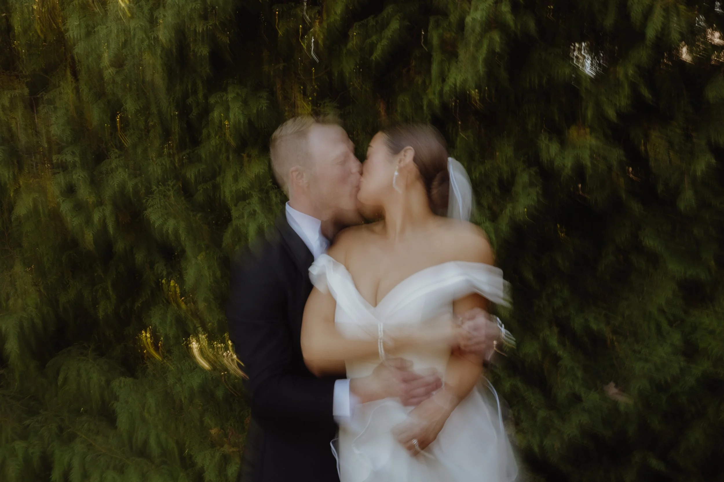 A bride and groom share a kiss, standing in front of a leafy green background, with motion blur around them.