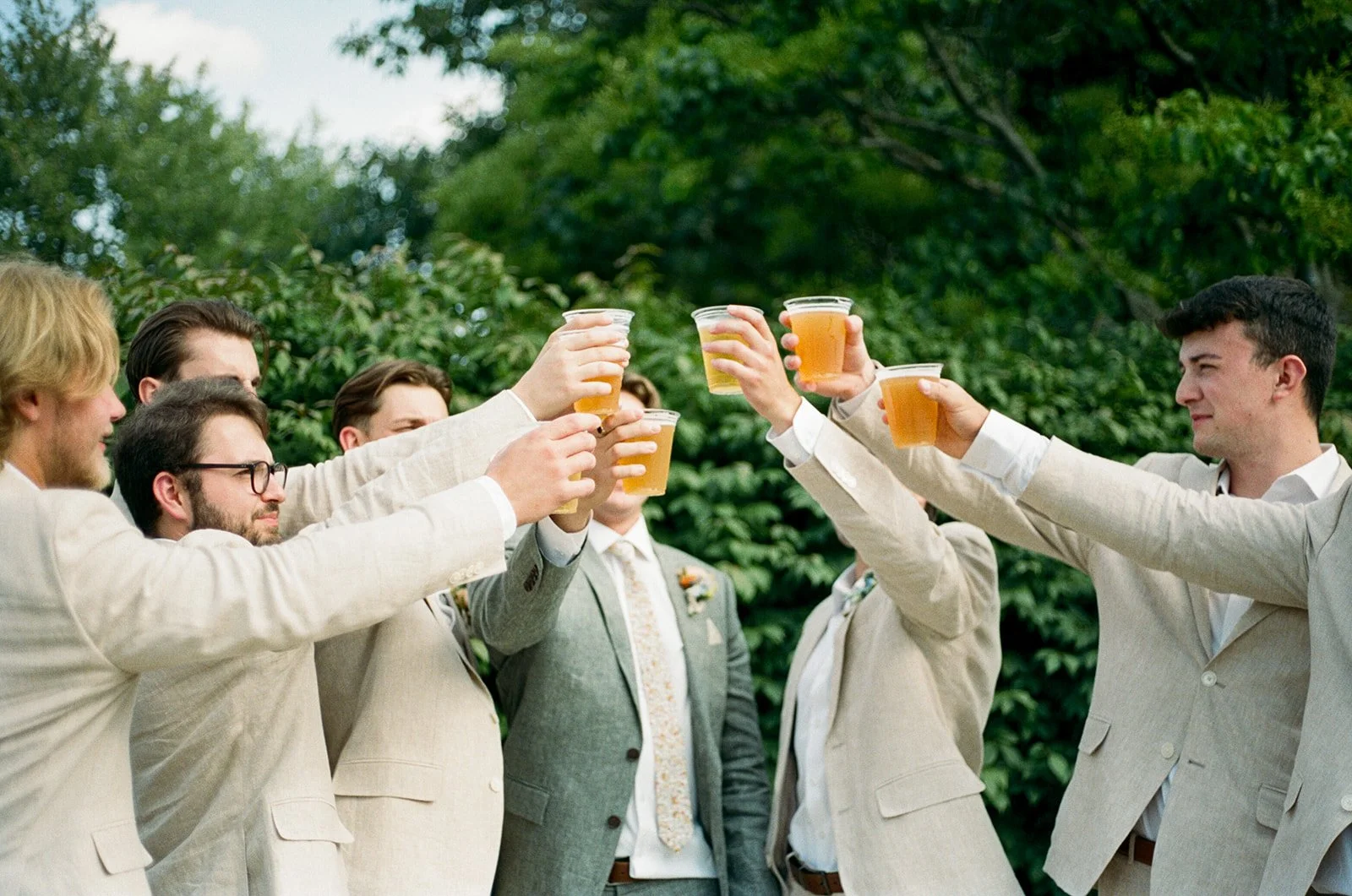Group of men in suits raising glasses of beer in a toast outdoors surrounded by greenery.