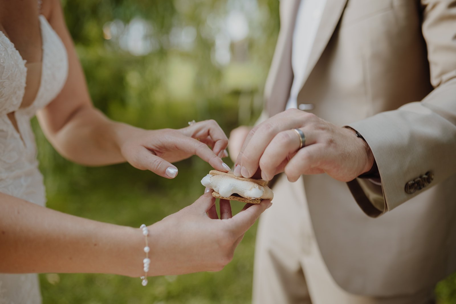 A couple's hands exchanging a cookie with white cream filling during an outdoor wedding ceremony.