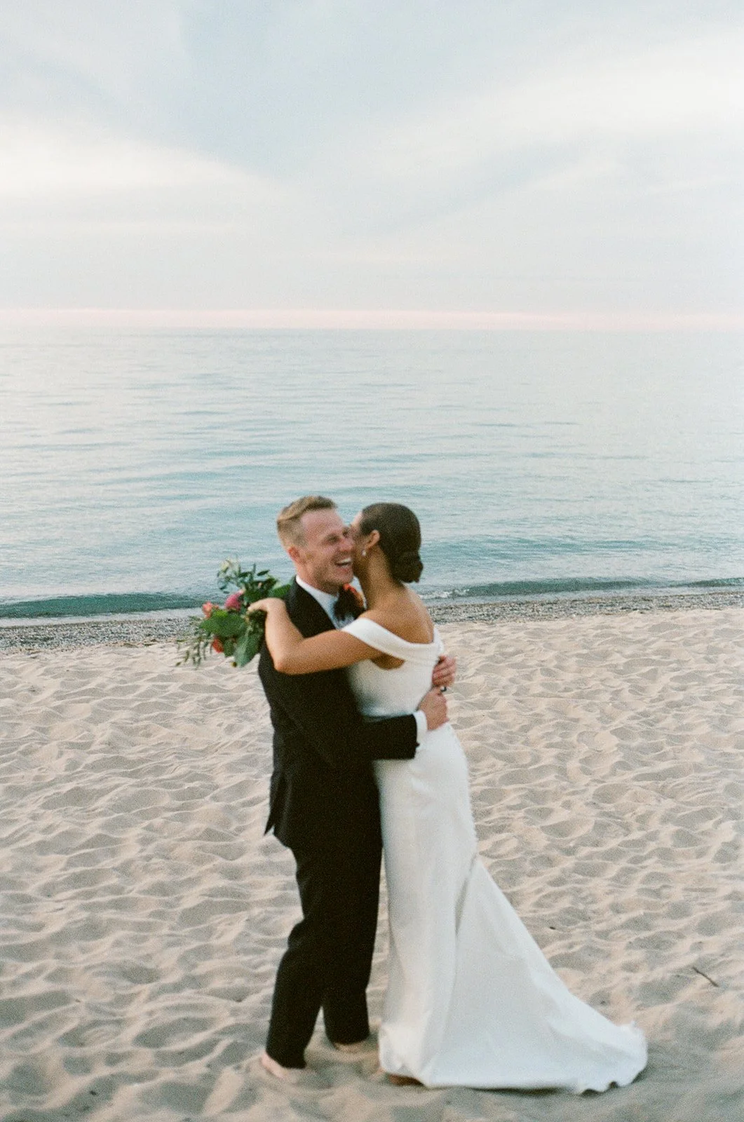 A bride and groom hugging on the beach at sunset, with the ocean in the background.