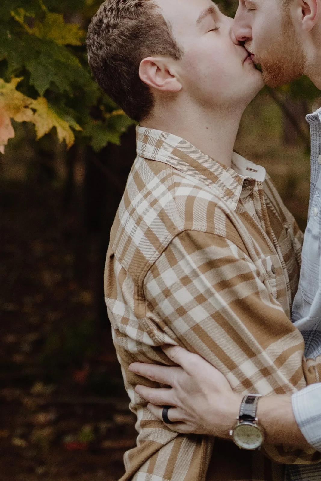 Two men kissing outdoors, surrounded by autumn leaves, embracing each other.