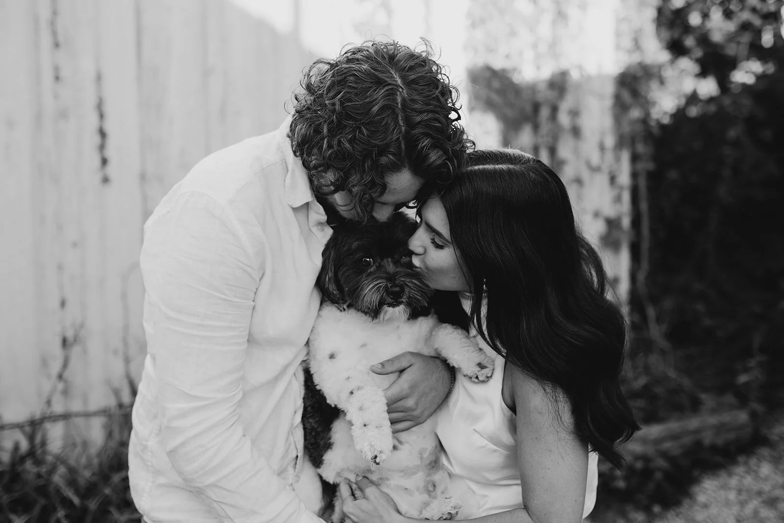 A black-and-white photograph of a couple holding a small puppy together outdoors, with all three leaning in affectionately, touching noses, and the woman kissing the puppy's face.