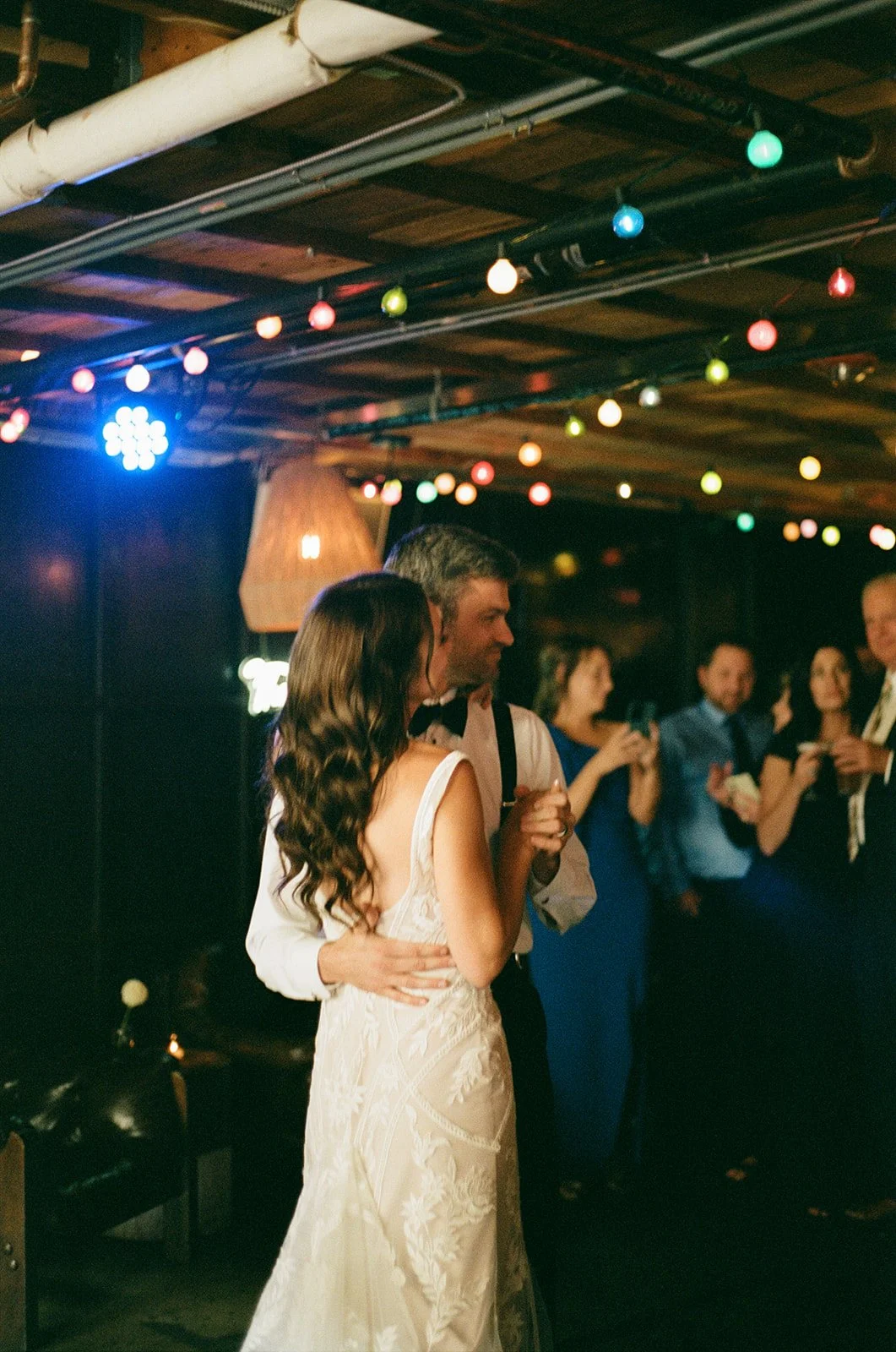 A bride and groom dancing closely at their wedding reception, surrounded by guests in a dimly lit venue with colorful string lights overhead.