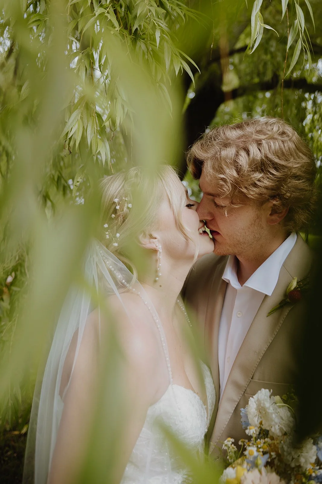 A bride and groom share a kiss in a lush, green outdoor setting, partially obscured by leaves.