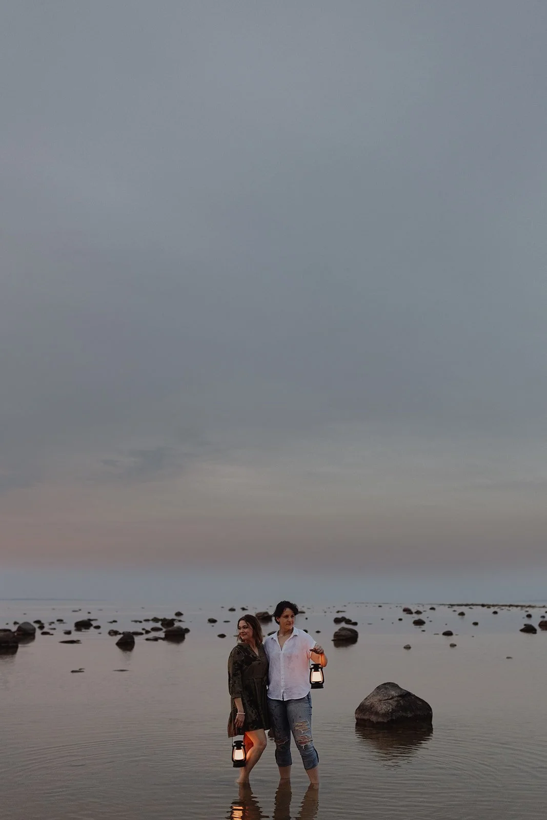Two women standing in shallow water on a beach at dusk, holding lanterns, with rocks in the background and a cloudy sky.