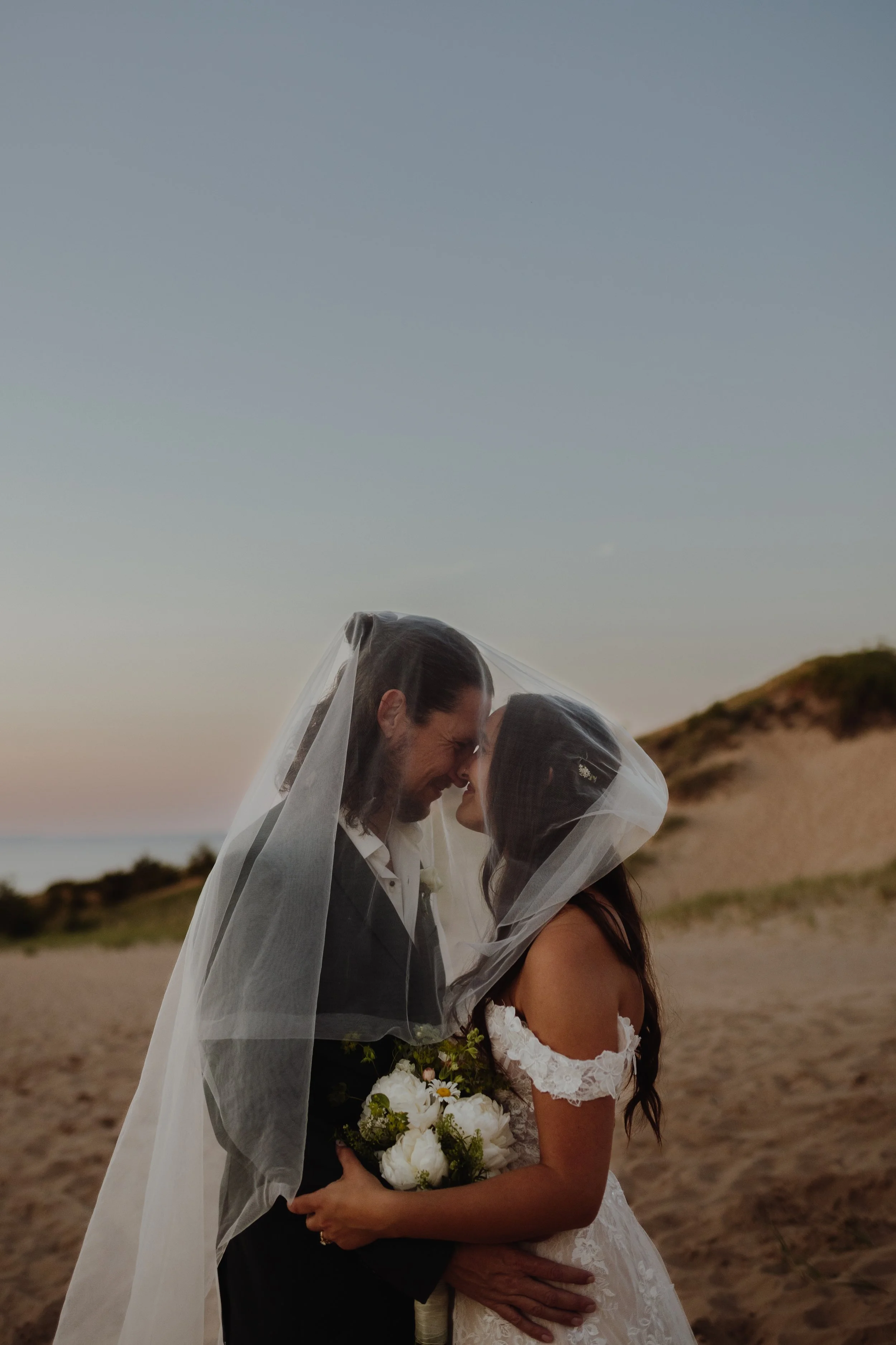 A newlywed couple stands on the beach, sharing a private moment under a sheer veil at sunset.