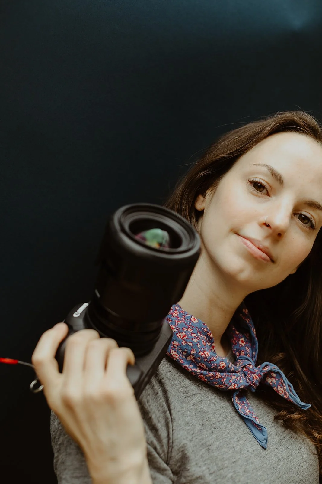 A woman with brown hair, wearing a light gray top and a patterned blue scarf, holding a camera with a large lens, standing against a black background.