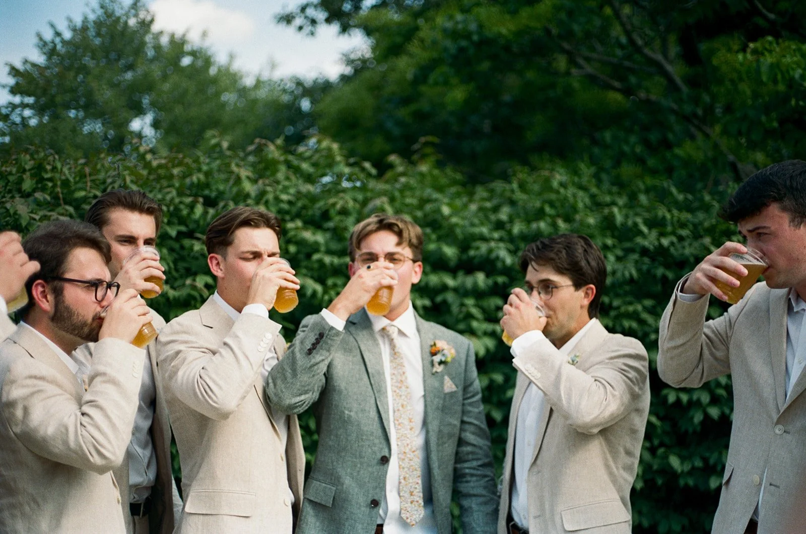 Group of six men dressed in suits drinking beer outdoors with green bushes and trees in the background.