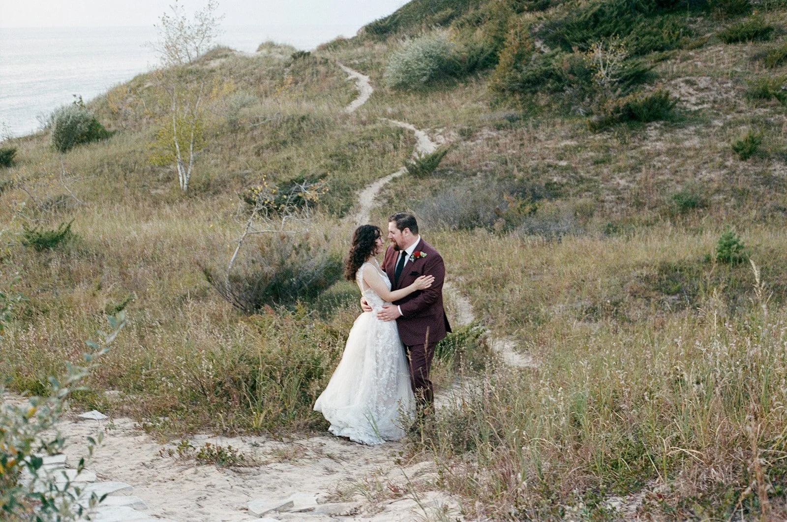 A bride and groom embrace on a sandy trail in a grassy, hillside landscape with a body of water in the background.