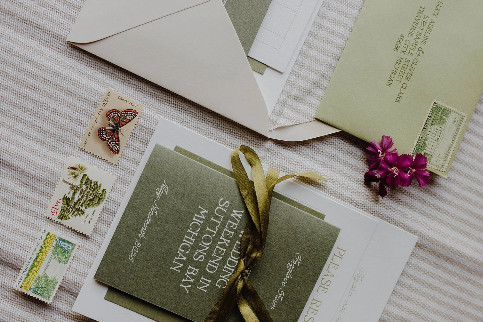 Wedding invitation card with green and white paper, tied with a gold ribbon, next to pink flowers, surrounded by postage stamps and an envelope on a striped fabric surface.
