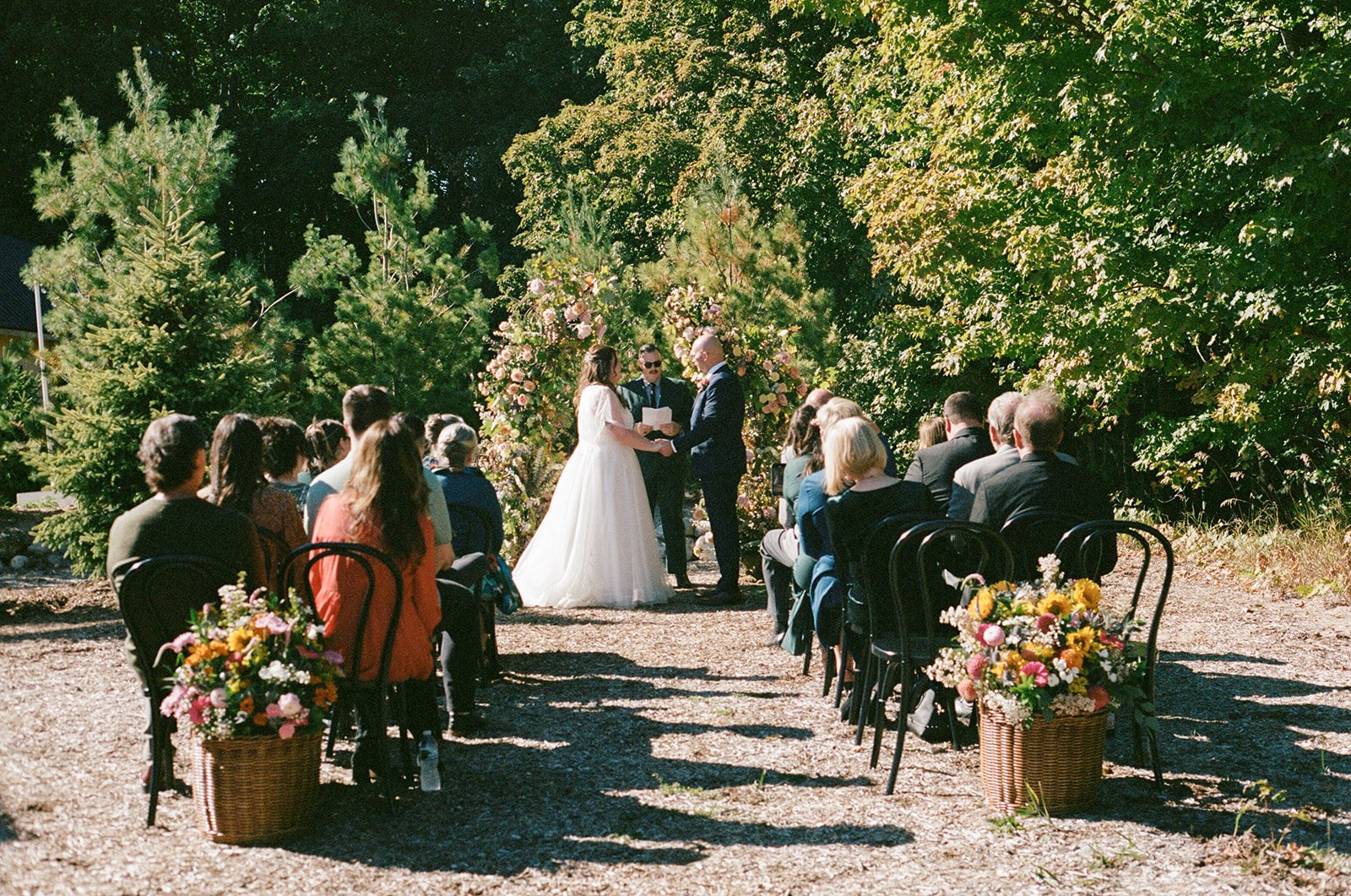 A wedding ceremony outdoors with a bride and groom standing before an officiant, surrounded by guests sitting on black chairs and floral arrangements, with green trees in the background.
