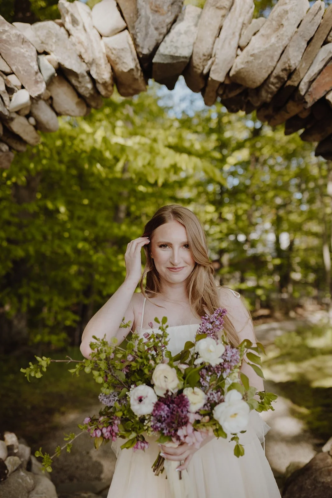 Woman holding a bouquet of white and purple flowers outdoors, standing under a stone archway with green trees in the background.