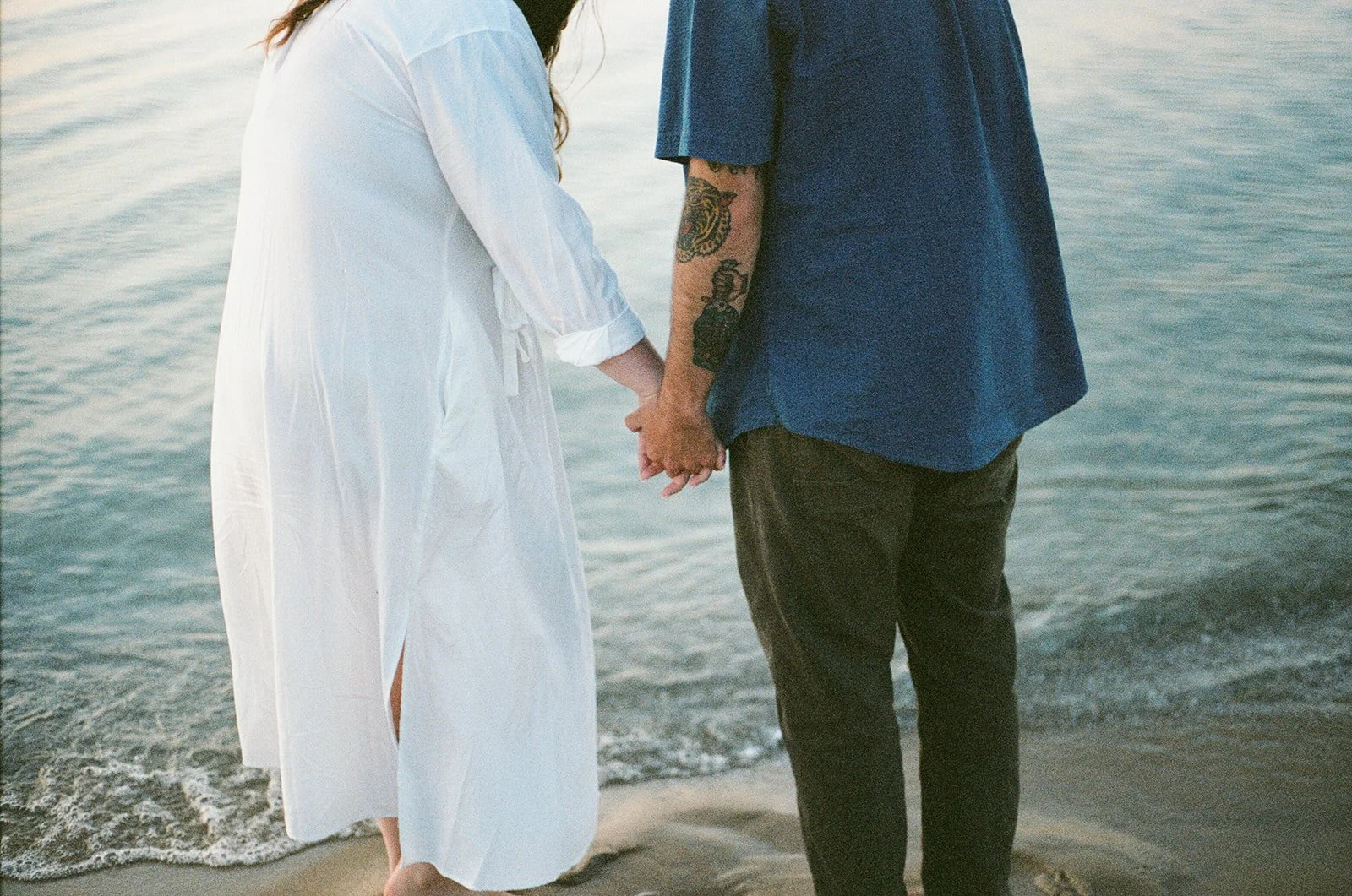 A couple holding hands and standing on the beach near the water, seen from behind.