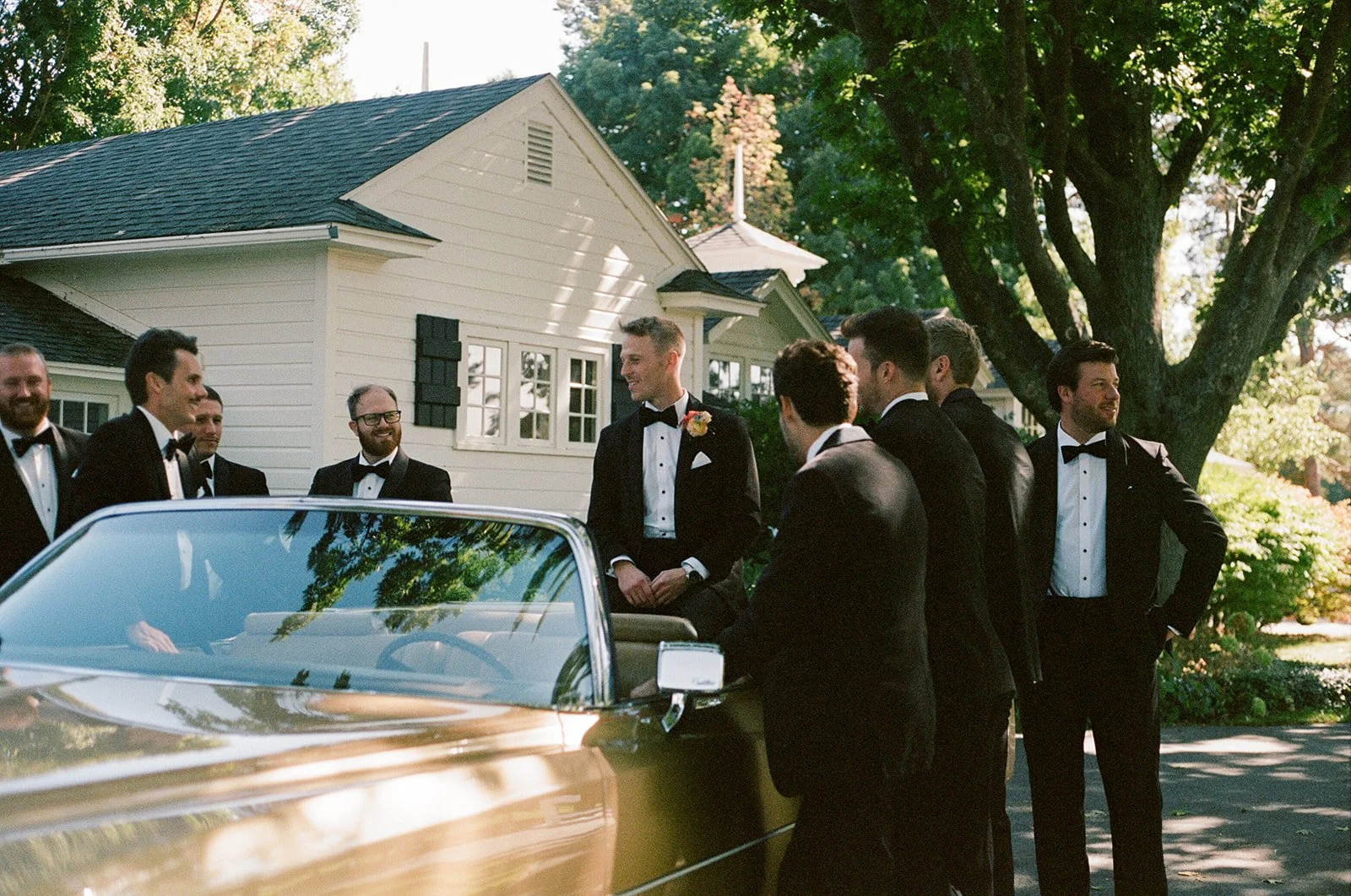 A group of men dressed in black tuxedos, some with bow ties, standing near a vintage gold convertible car in front of a white house with a porch, large trees, and green foliage, enjoying a sunny day.