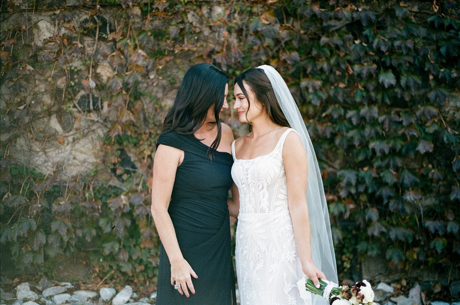 A bride in a white wedding dress and veil standing close to a woman in a black dress, both with foreheads touching, outdoors against a background of dark green ivy-covered wall.