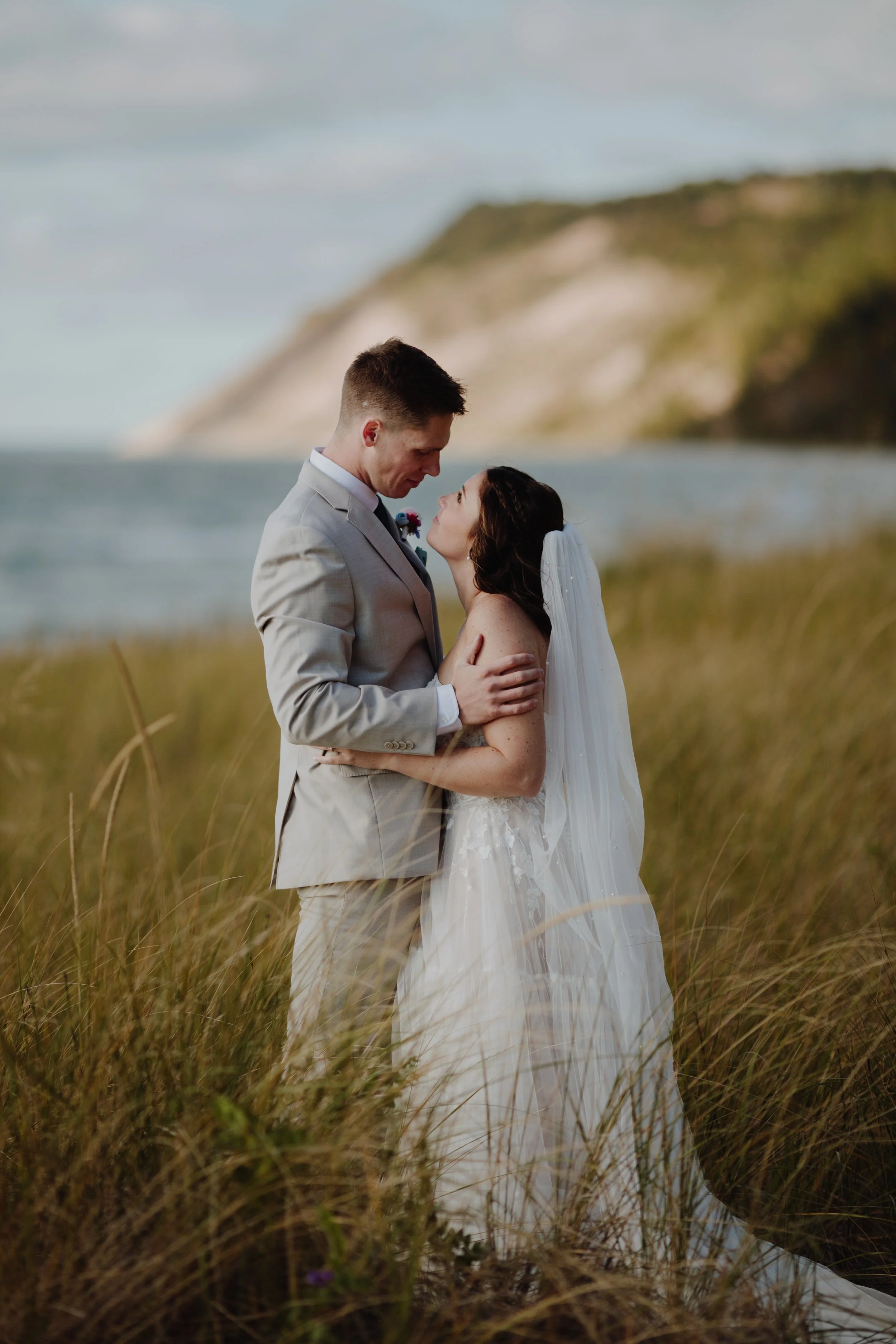 A bride and groom embrace on a grassy beach with a lake and cliffs in the background, during their wedding photoshoot.
