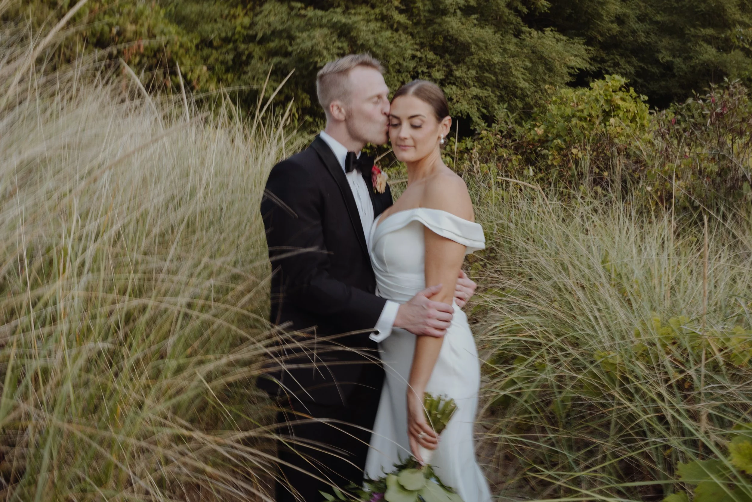 A bride and groom in wedding attire embrace and share a kiss outdoors among tall grass and greenery, with a backdrop of shrubs and trees.