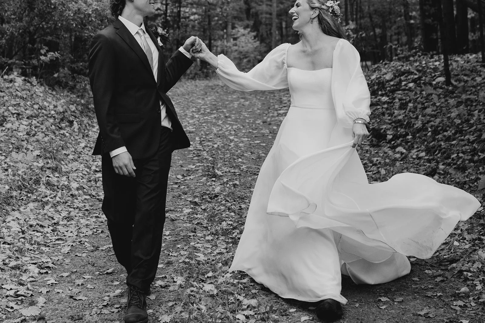Black and white photo of a bride and groom walking in a wooded area, holding hands and smiling.