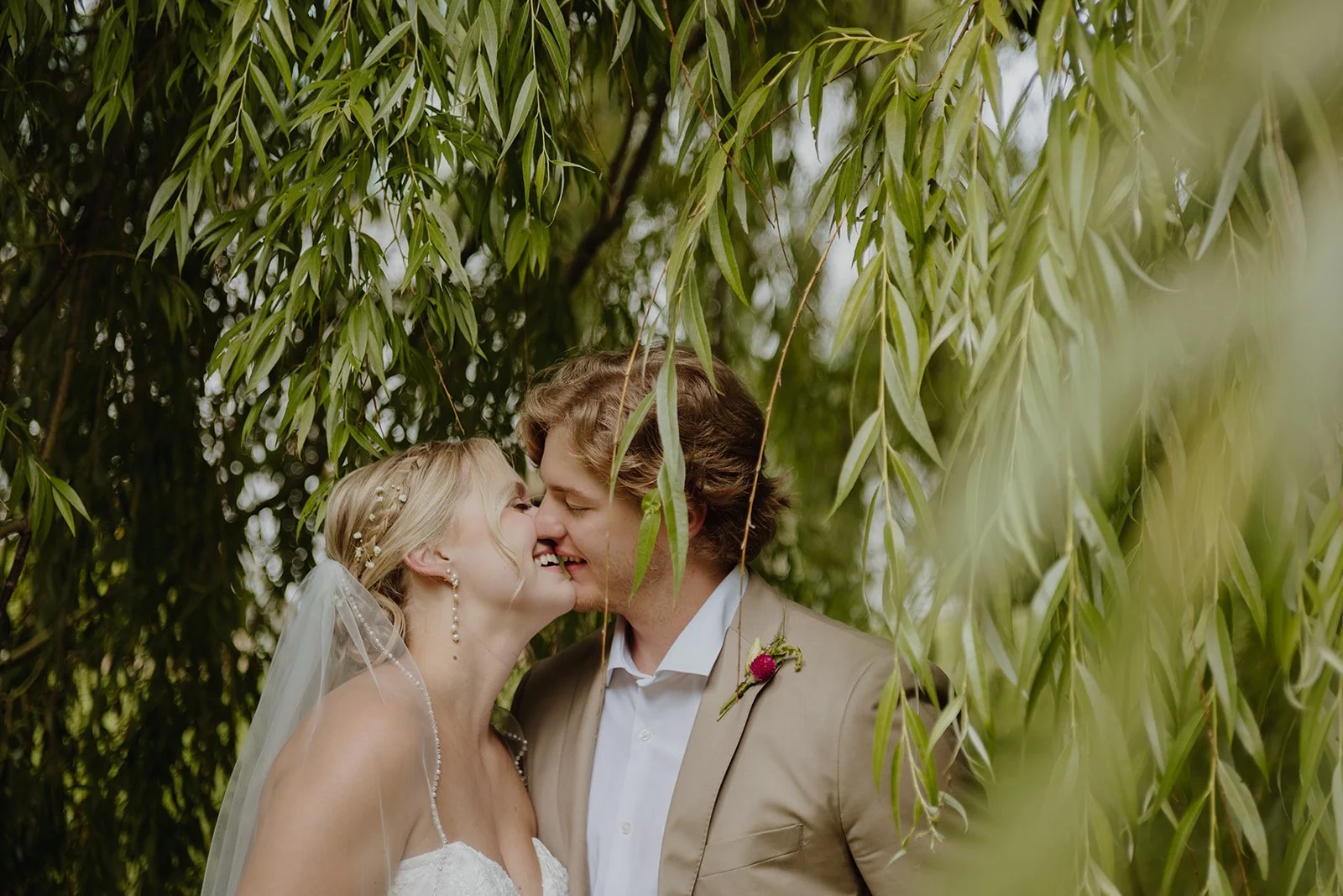 A bride and groom share a kiss in an outdoor setting surrounded by green foliage.