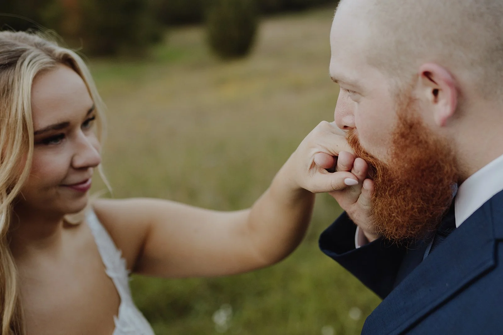 A woman gently touching a man's beard as he kisses her hand outdoors.