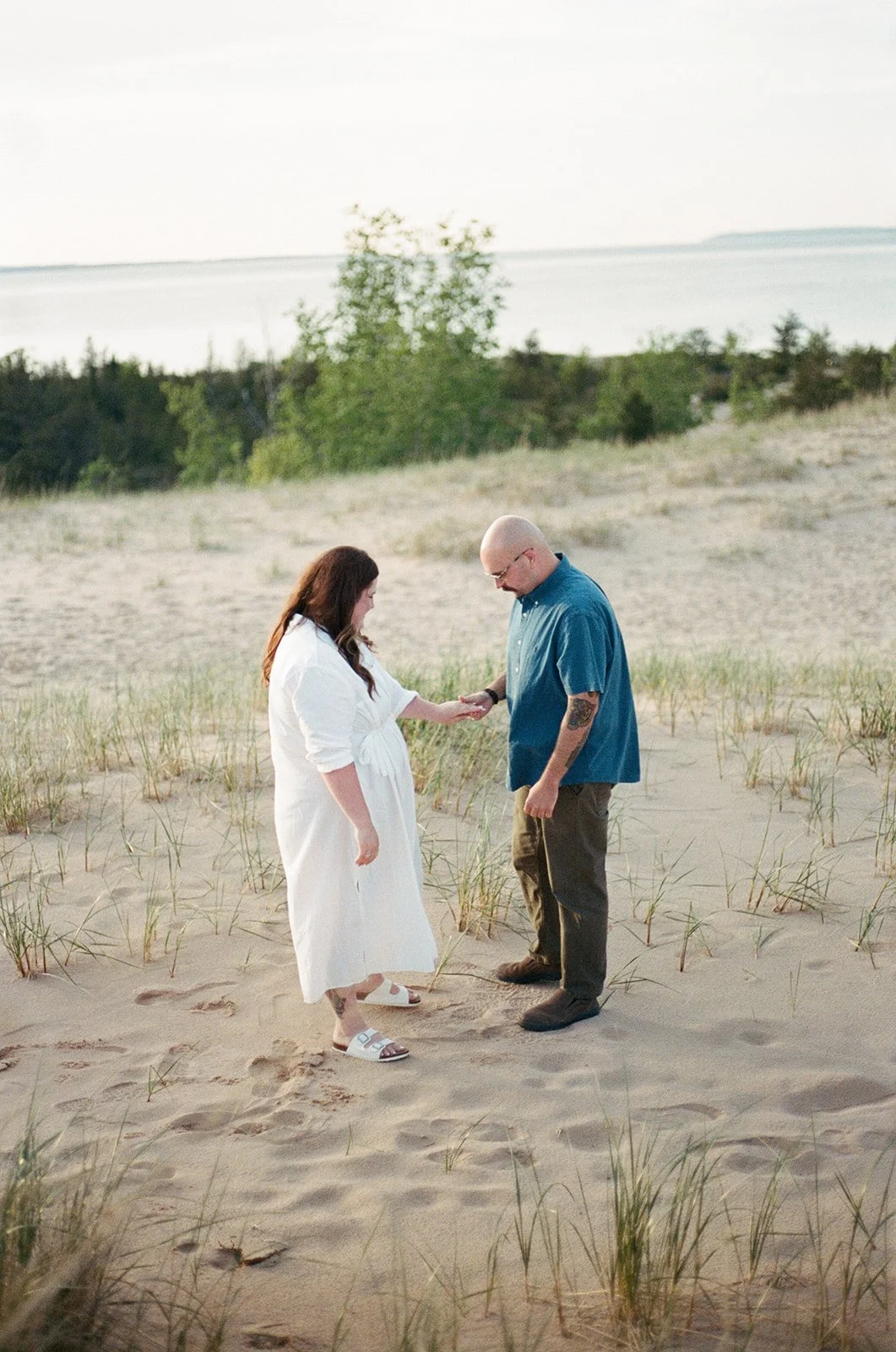 A couple standing on a sandy beach exchanging rings during their wedding ceremony, with the ocean and trees in the background.
