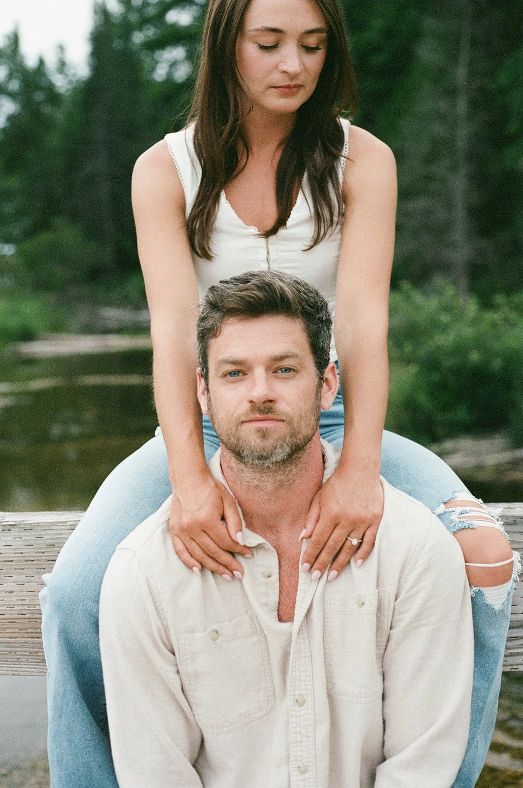 A woman sits on a man's shoulders by a lake, with the woman placing her hands on the man's shoulders. They are outdoors with trees and water in the background.