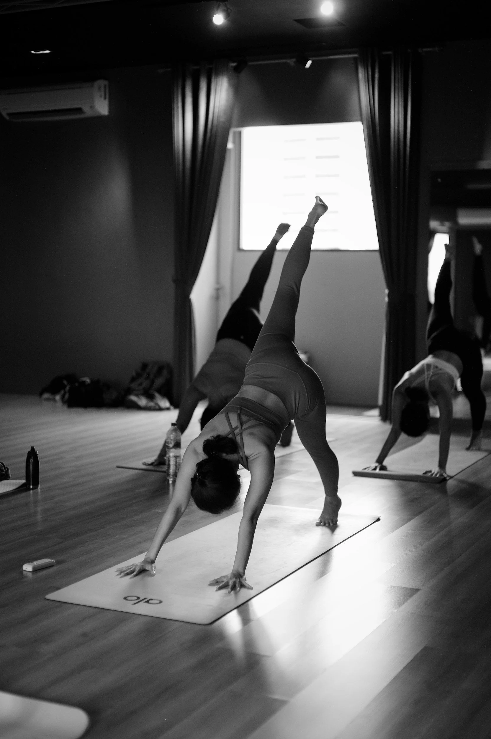 People participating in a yoga or fitness class, performing handstand poses on yoga mats in a room with dark curtains and a window.