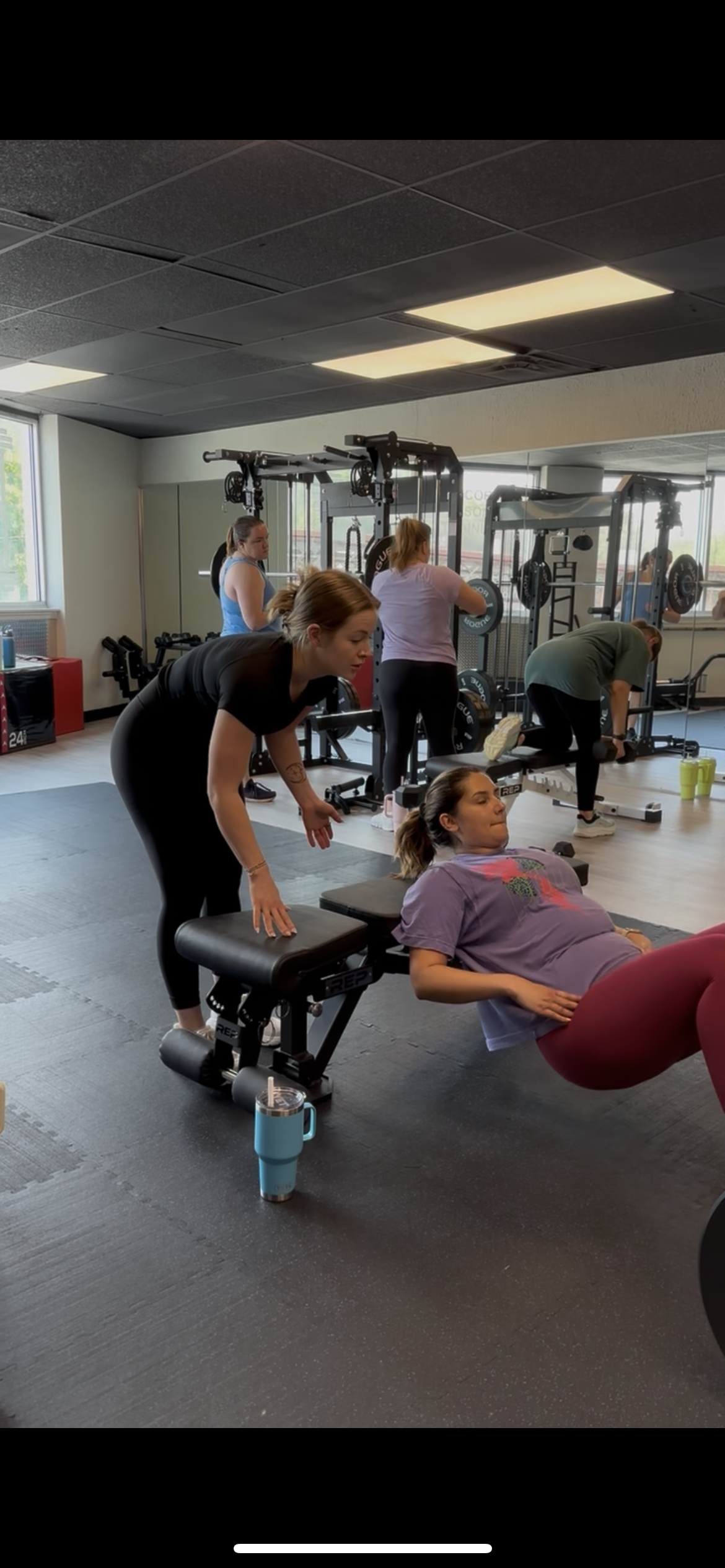 Women working out at a gym, with one woman doing a glute bridge exercise on a bench and others lifting weights in the background.