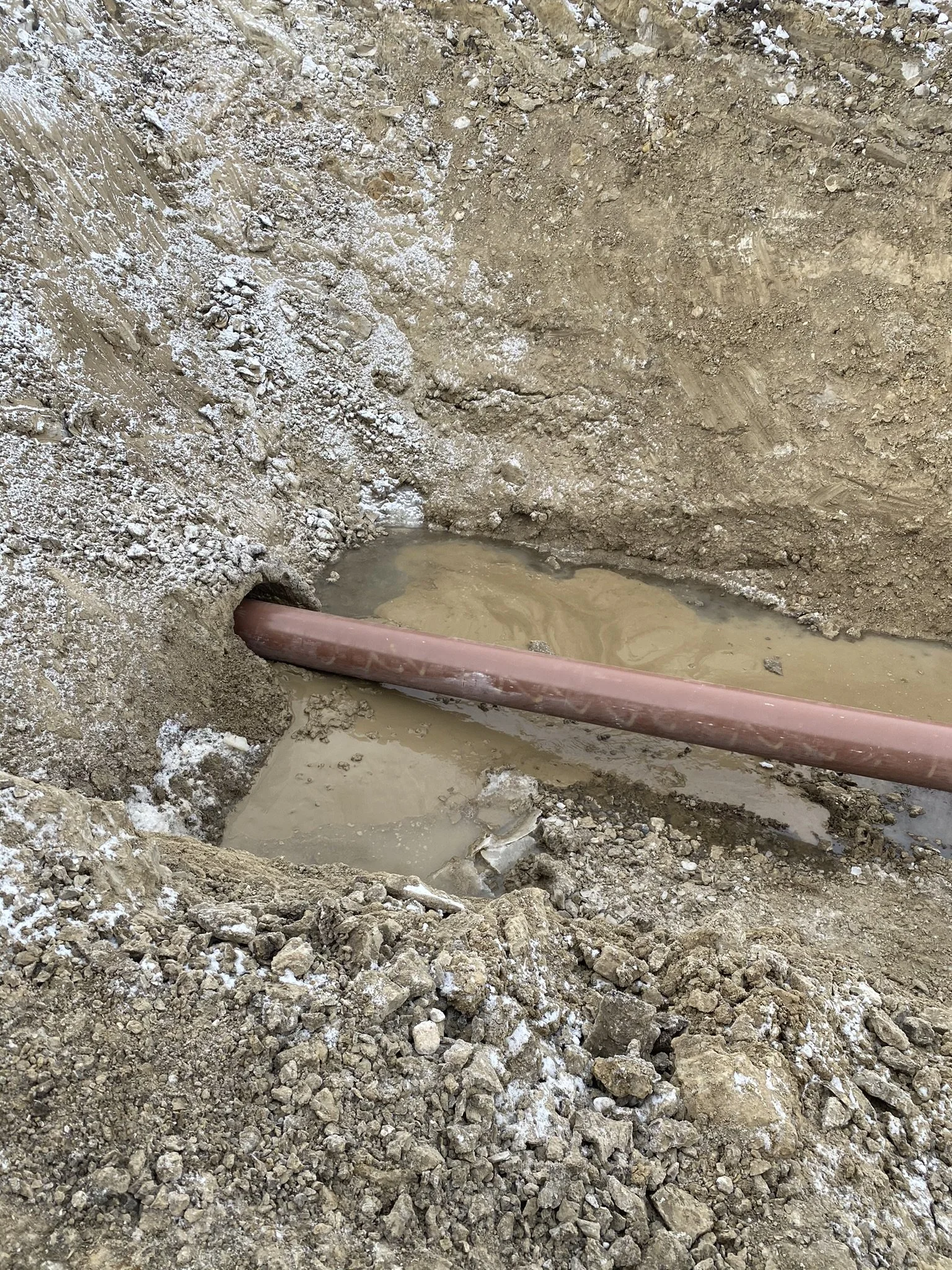 Underground construction site showing a colored pipe running through a hole in the dirt, with muddy water collected at the bottom of the excavation.