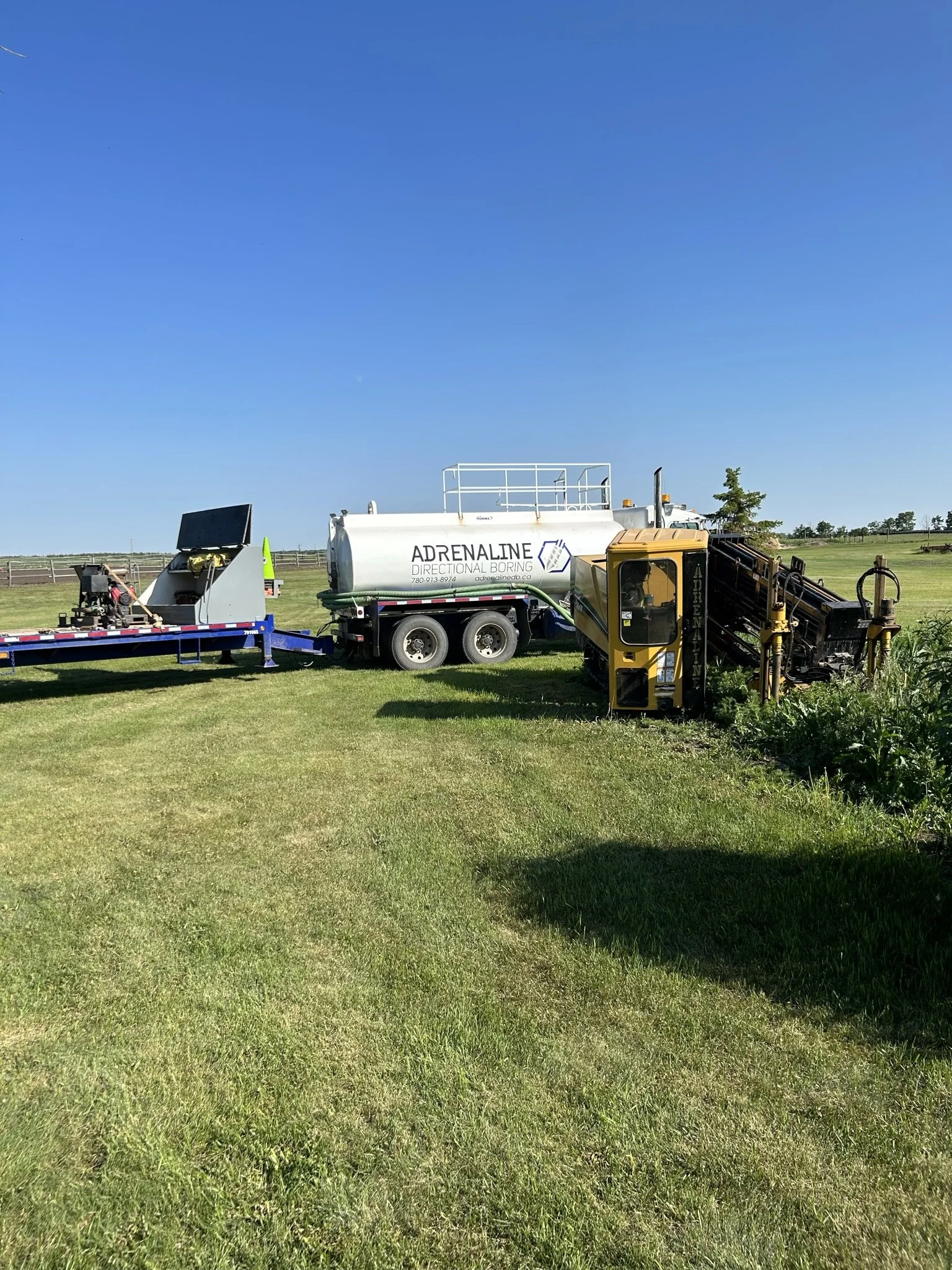 A dirt drilling rig and a tanker truck labeled "Adrenaline" on a grassy field under a clear blue sky.
