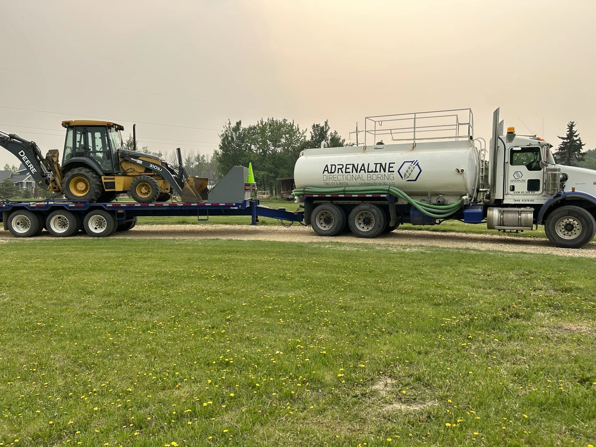 A large white tanker truck labeled 'ADRENALINE DIRECTIONAL BORING' is parked on a grassy area. The truck is pulling a flatbed trailer carrying a compact yellow John Deere backhoe loader.