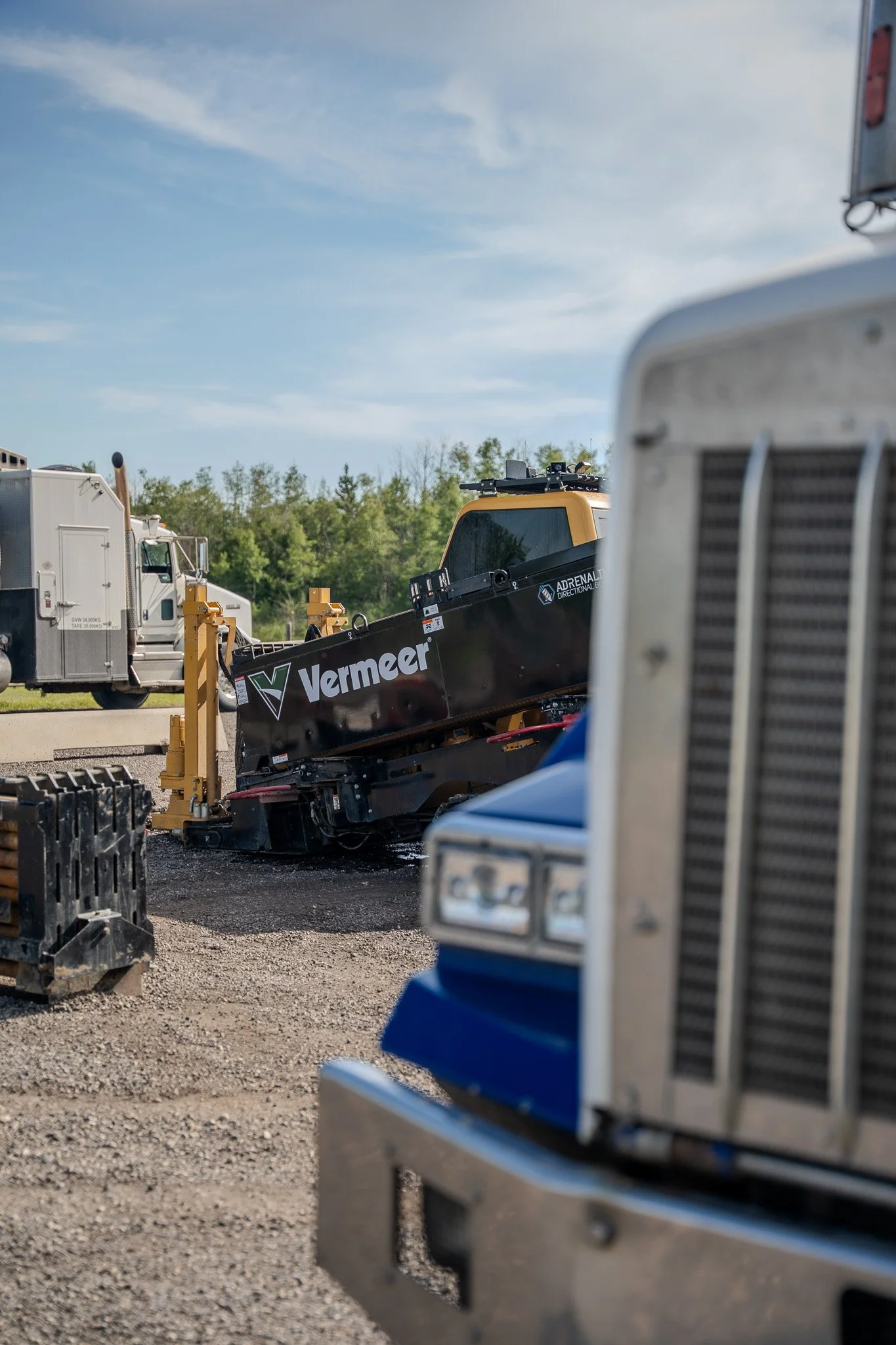 Construction equipment and vehicles parked outdoors on the dirt ground with trees and blue sky in the background.