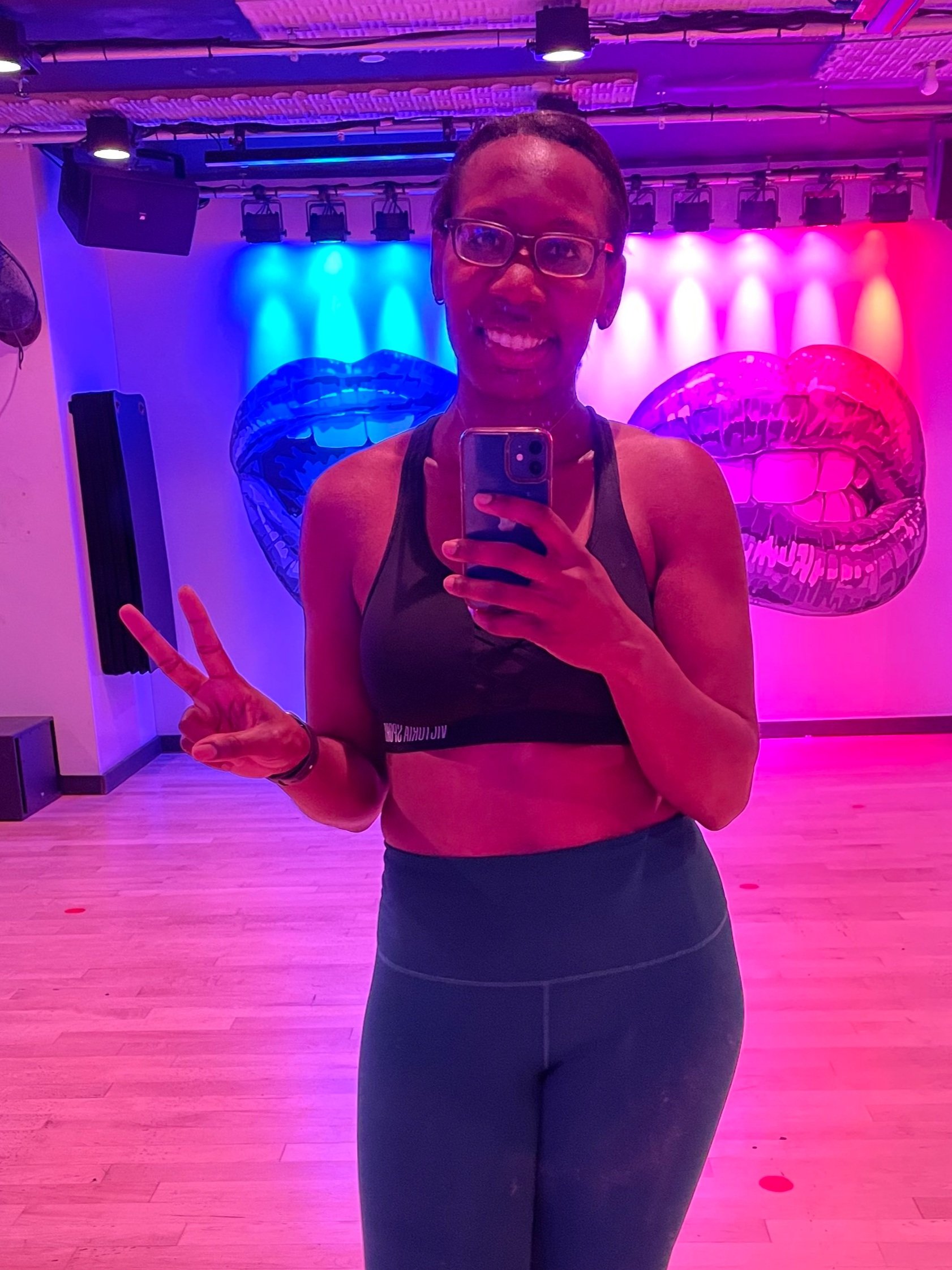 A smiling woman takes a post-workout mirror selfie in a colorful room with purple and pink neon lighting. 