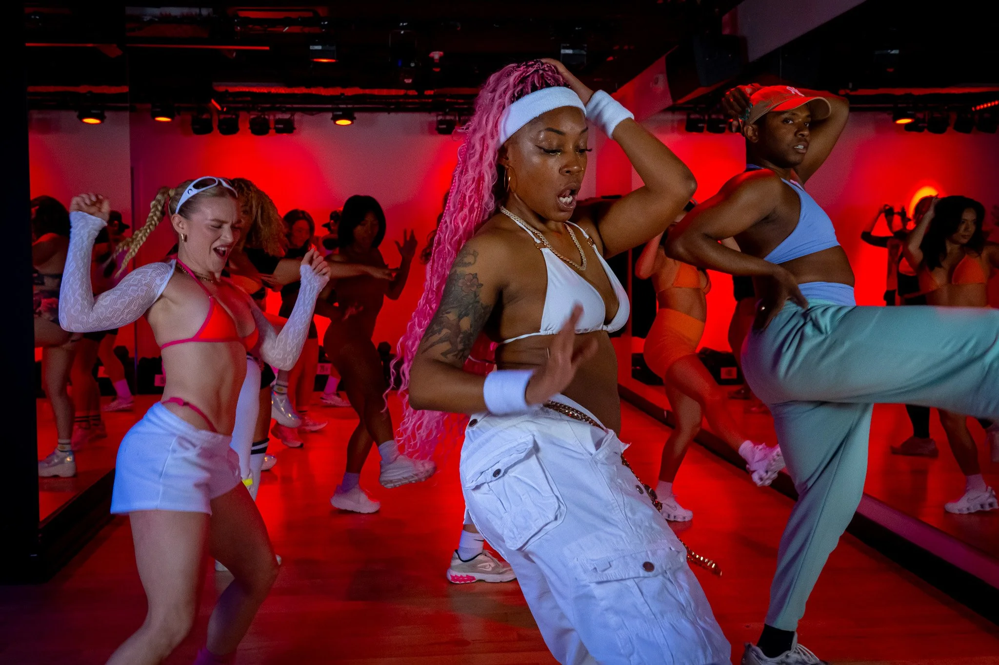 Group of diverse women dancing energetically in a dance studio with red lighting and mirrors.