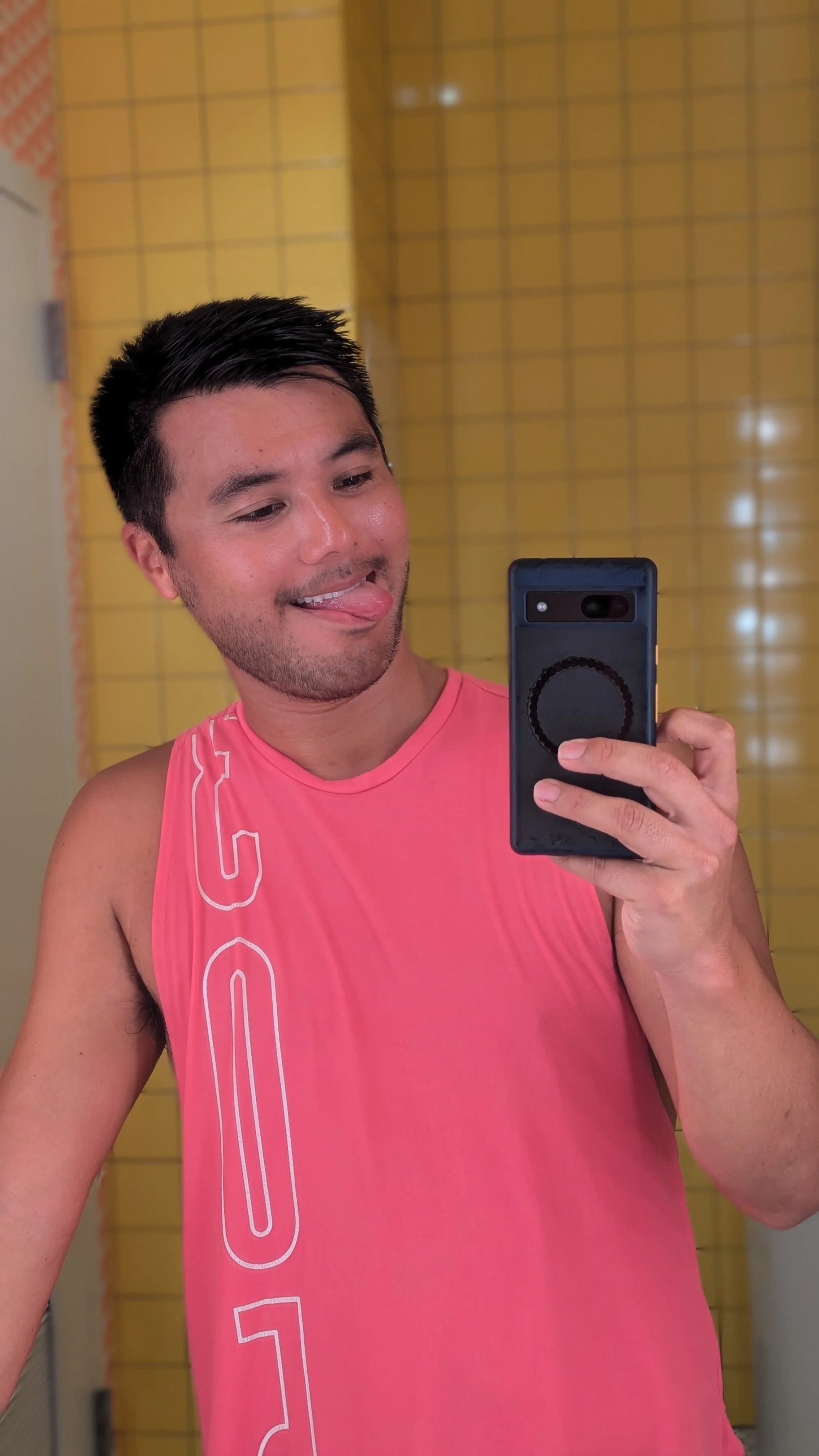 A young man taking a selfie in front of a yellow tiled wall, wearing a pink sleeveless athletic shirt, sticking out his tongue slightly.