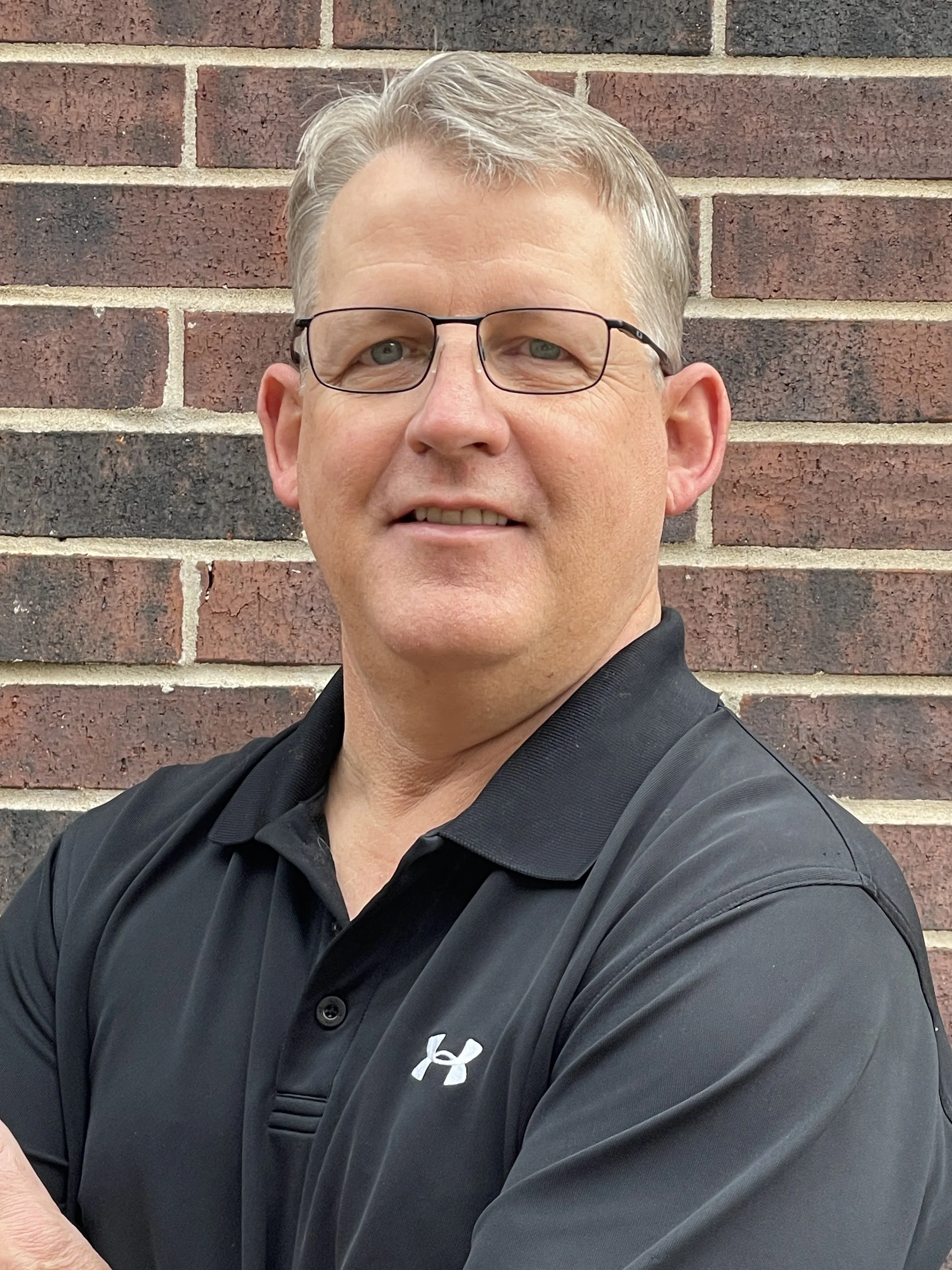 Man with glasses in a black collared shirt standing in front of a brick wall.
