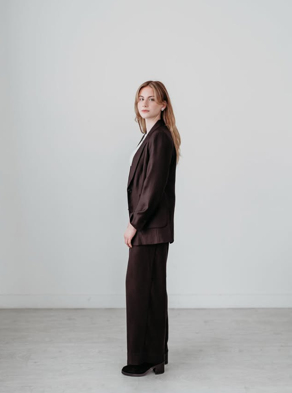 A woman with long blonde hair wearing a dark brown suit stands against a white wall in a minimalist room.