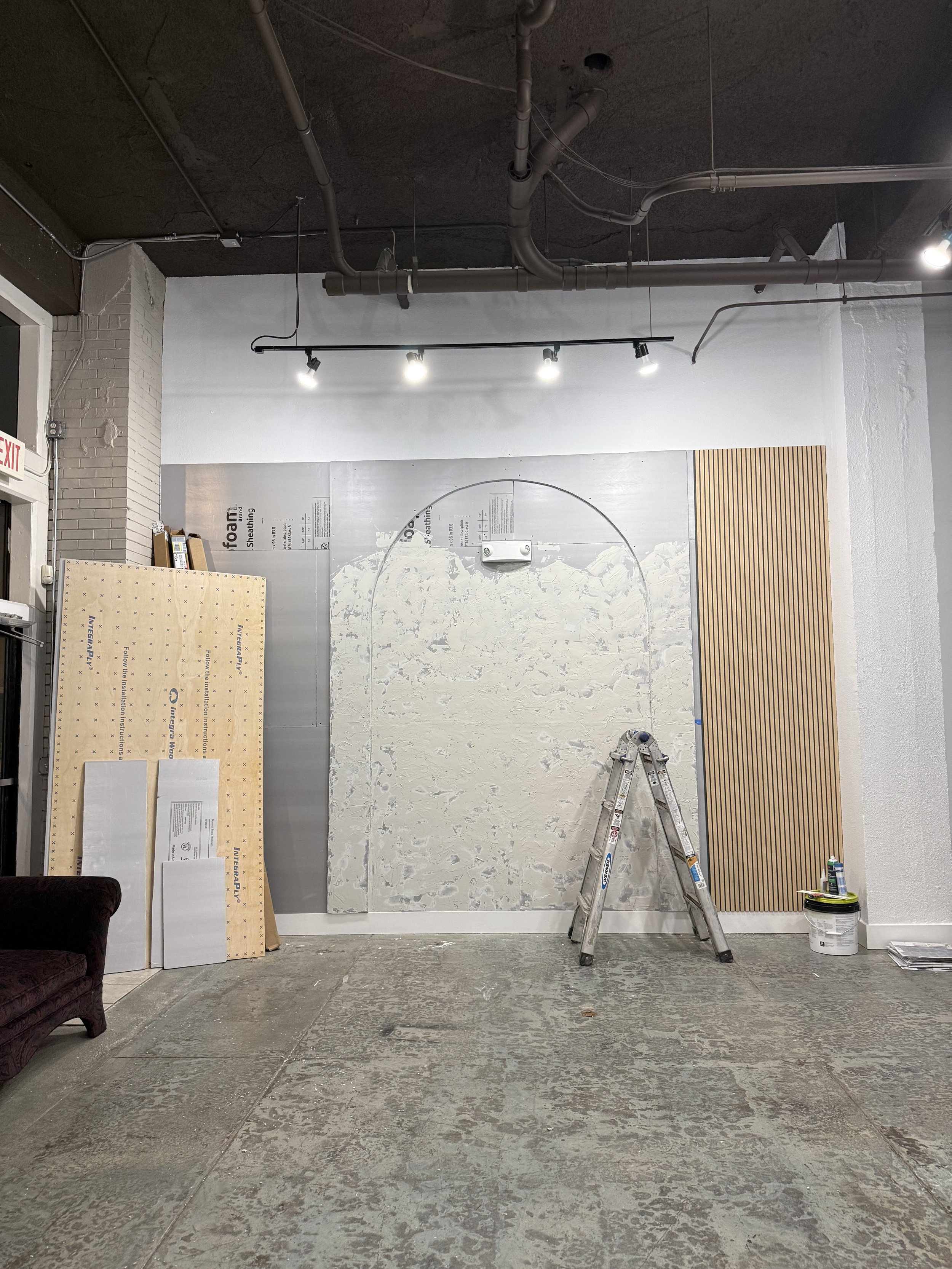 An indoor space under renovation with a variety of construction materials and tools. There are large sheets of drywall, a step ladder, a bucket, grout, and a decorative wooden panel on the wall. The floor and ceiling are unfinished, with exposed pipe