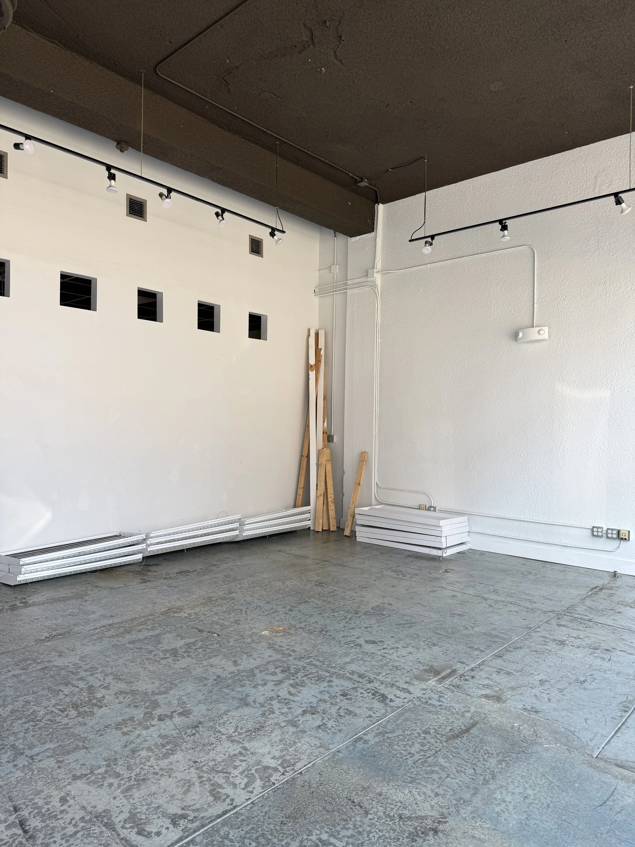Empty room with a concrete floor, white walls, some wooden planks, and rolled-up white boards or panels leaning against the wall. Overhead lighting and electrical outlets are visible.