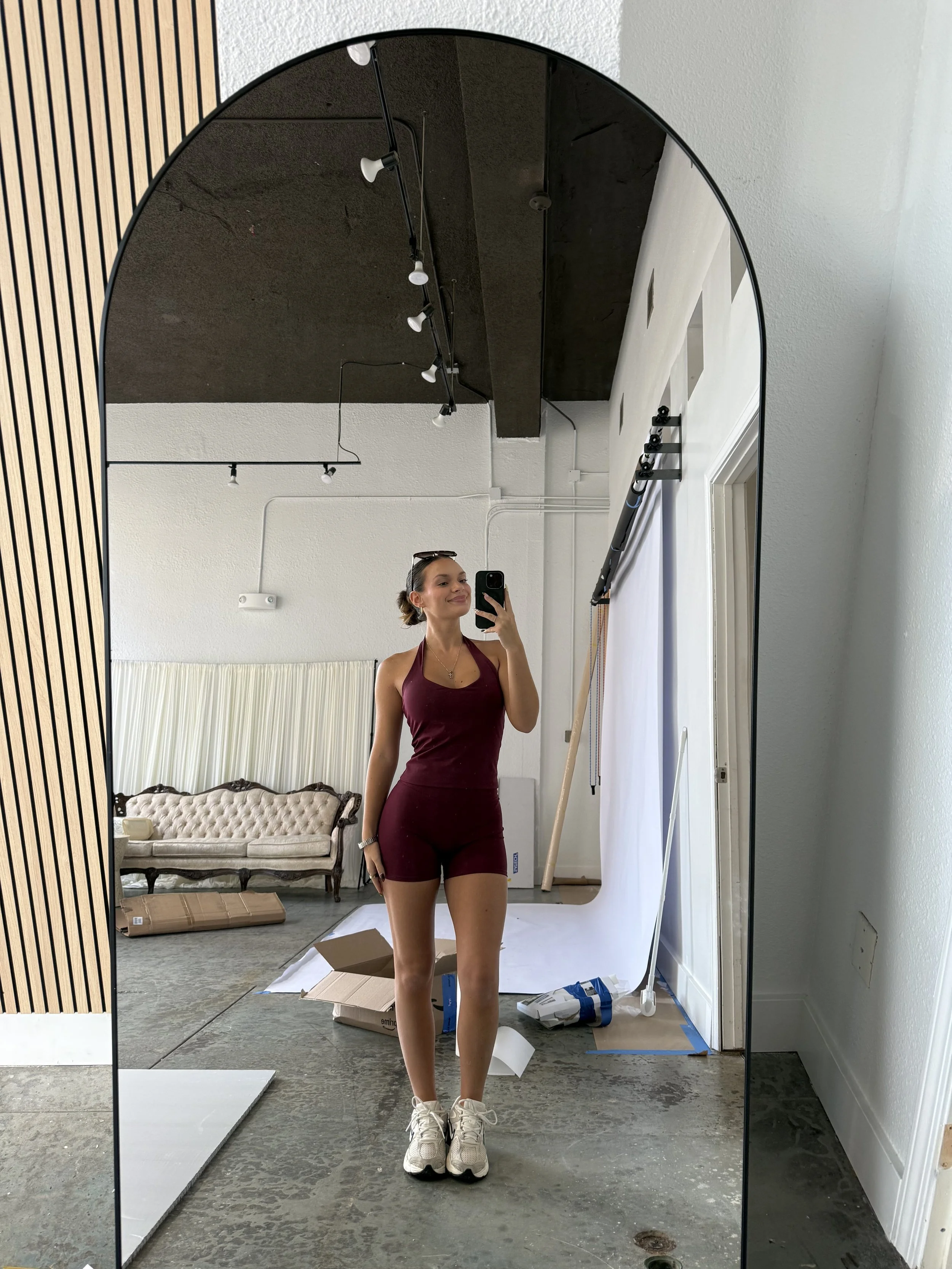 A woman in a burgundy workout outfit, wearing white sneakers, taking a selfie in a large, curved mirror in a room with some furniture and boxes on the floor.
