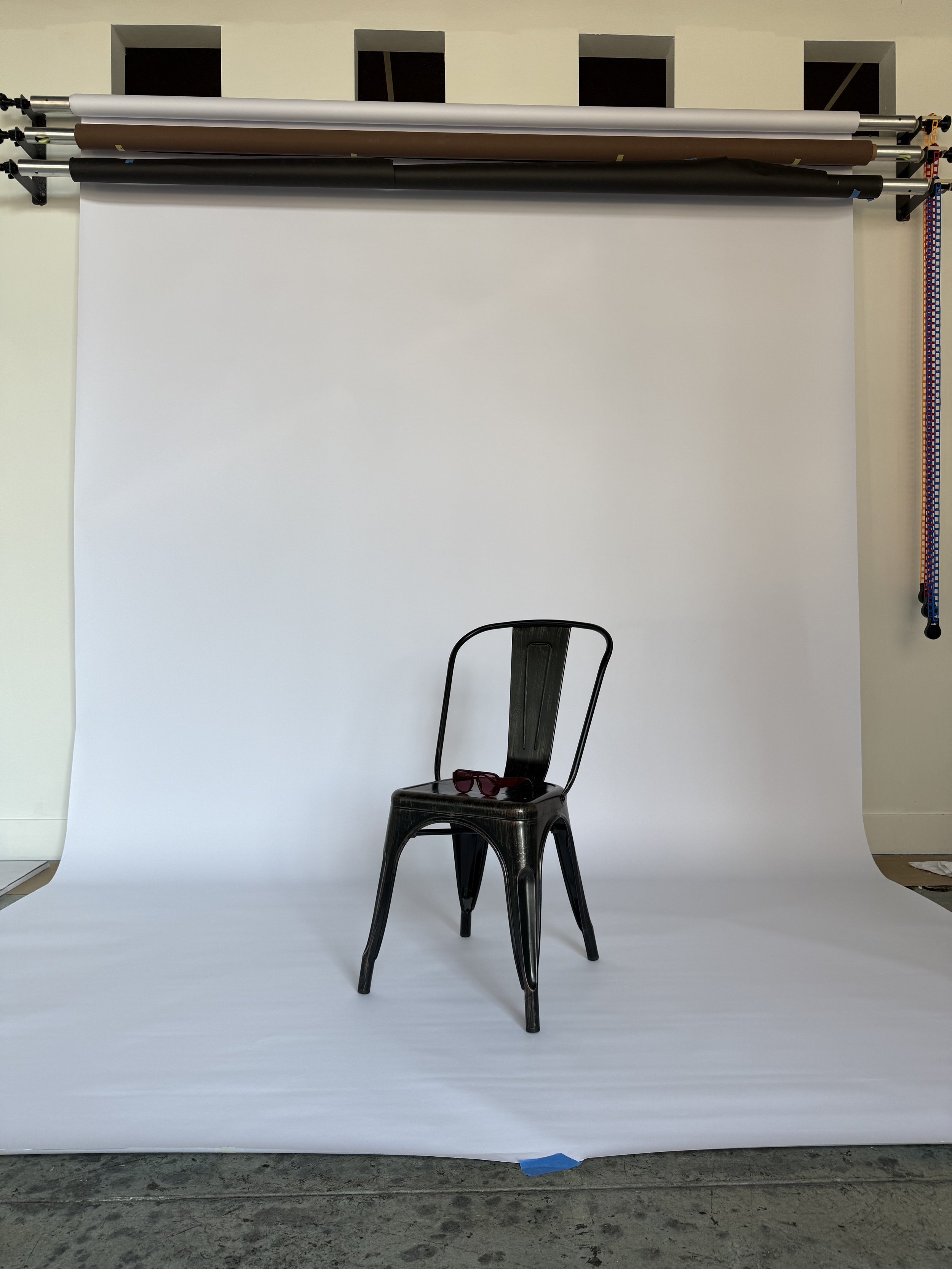 A black chair with a pair of sunglasses on its seat, in front of a white backdrop and backdrop stand with four rolls of background paper in gray, brown, black, and dark gray.