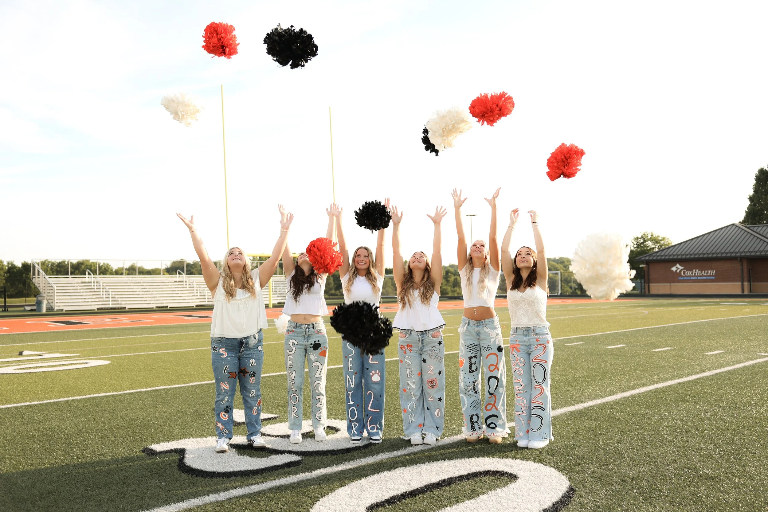 A man kneeling on a pink tulip field proposes to a woman who is jumping with joy, both smiling in a sunny outdoor setting with trees in the background.