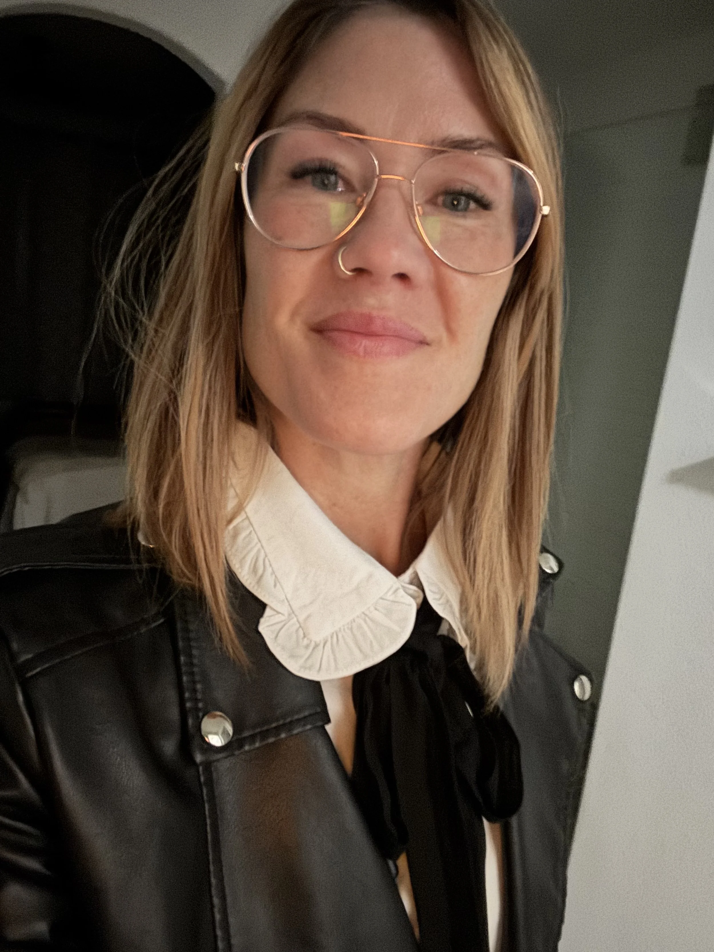 A woman with shoulder-length blonde hair, glasses, and a nose ring, smiling slightly, wearing a white ruffled collared shirt and a black leather jacket, in an indoor setting.