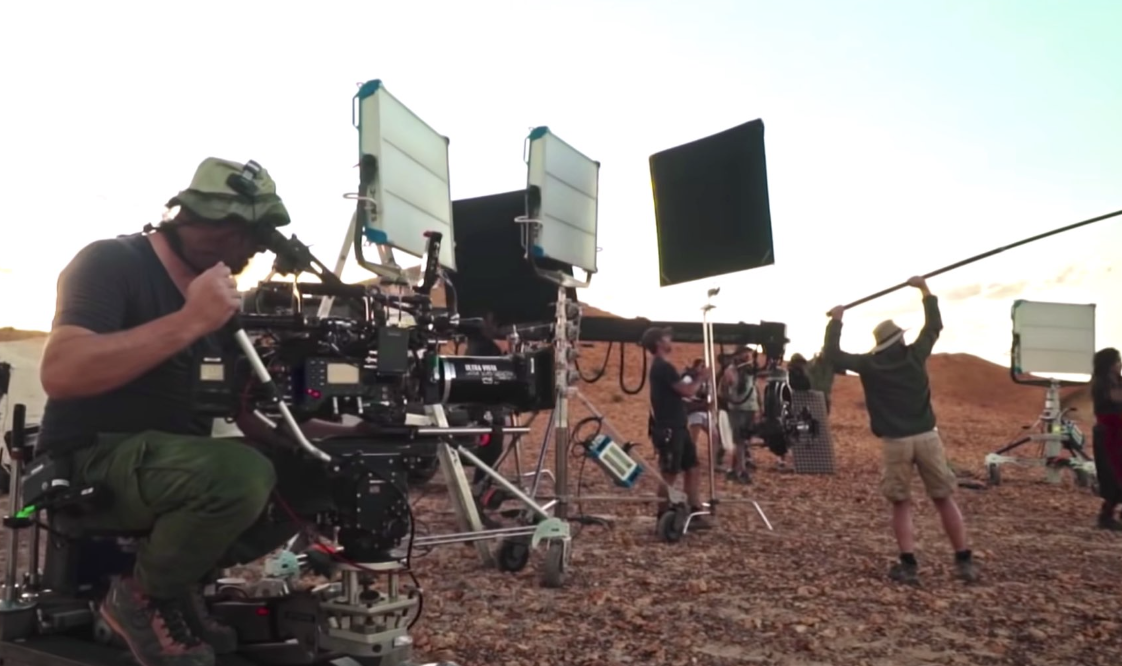 Film crew working outdoors on a desert landscape with large lights and monitor screens.