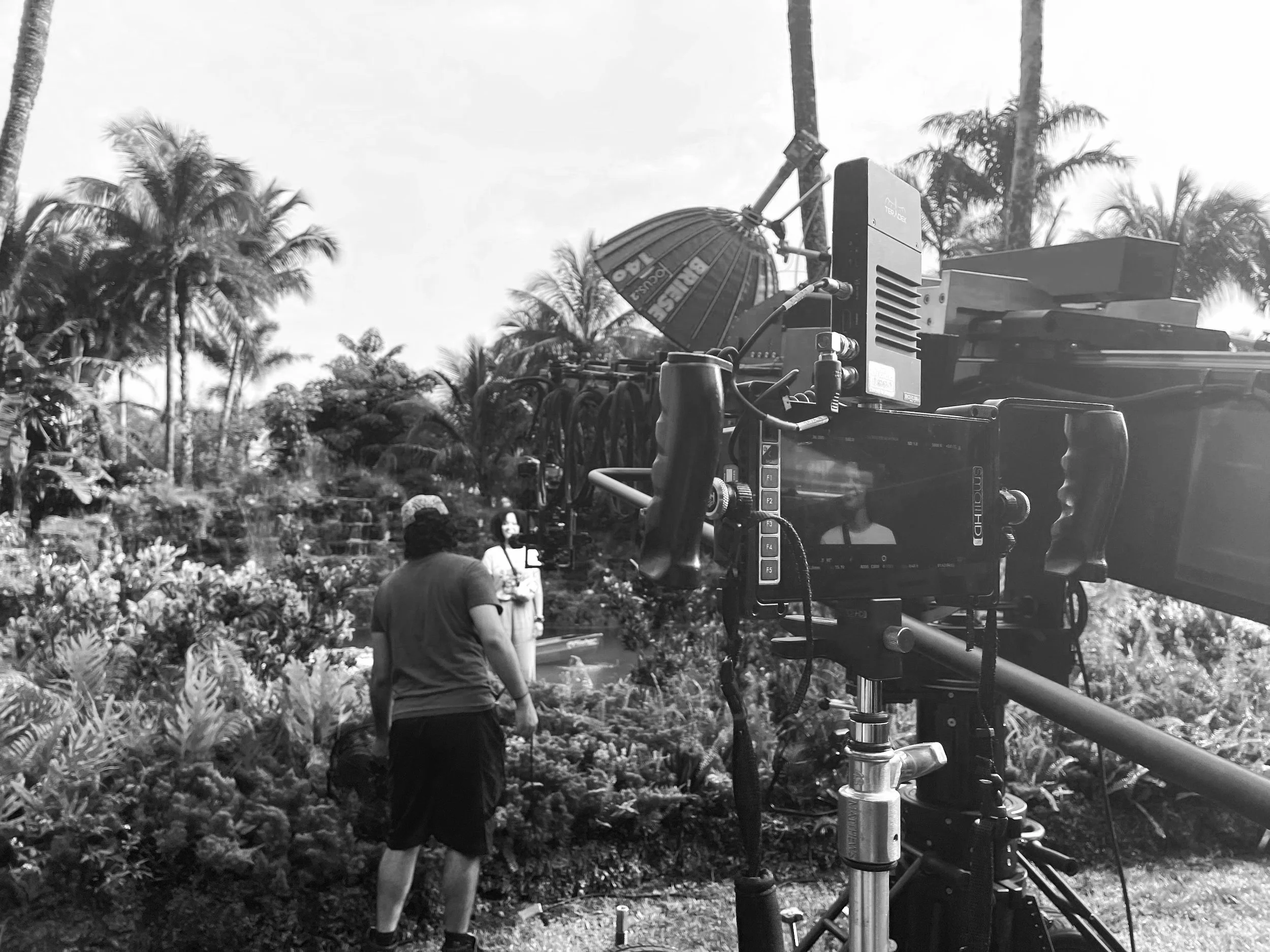 Black and white photo of a film crew filming outdoors with dirt, plants, and palm trees. Equipment visible includes a large camera, lights, and a red umbrella. Several people are present, one woman is talking or singing.