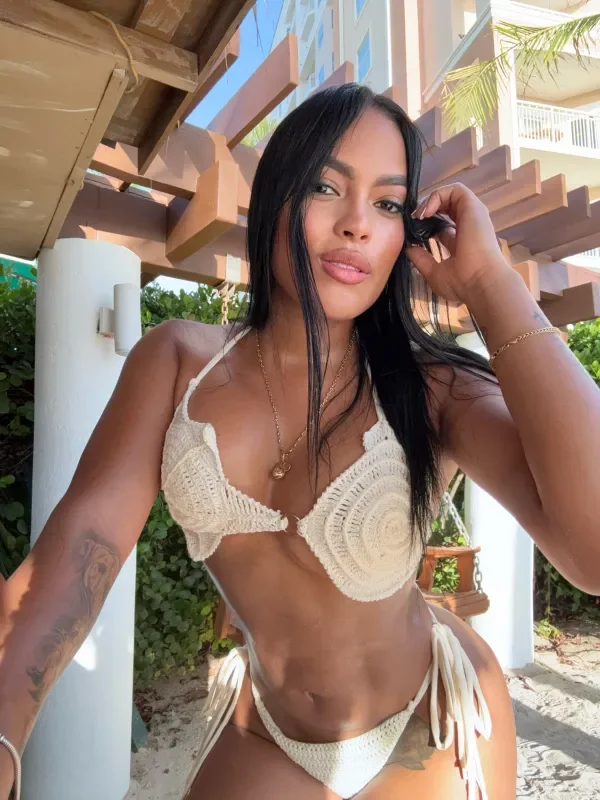 A woman in a crochet bikini taking a selfie outdoors with a wooden structure and lush greenery in the background.