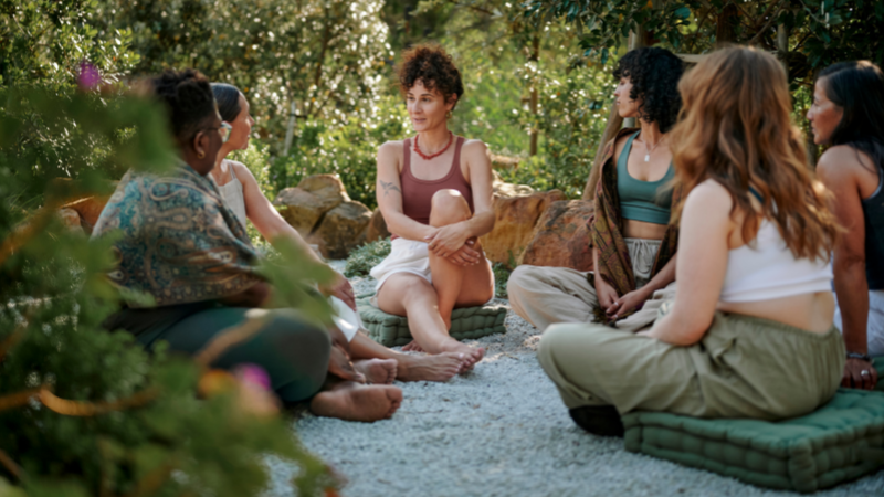 A circle of women chat in relaxed poses on meditation cushions, outdoors, surrounded by lush gardens