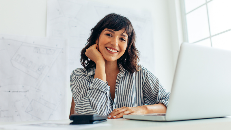 A woman architect at her laptop, with plans on the wall behind. Soft light spills in from the window to her right